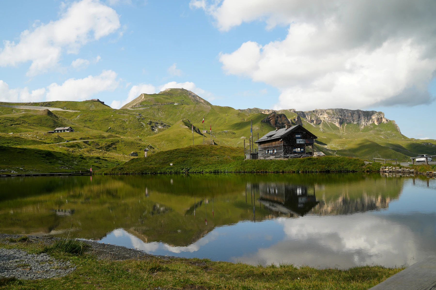 A wooden mountain cabin reflected in a calm alpine lake beneath green hills and clouds