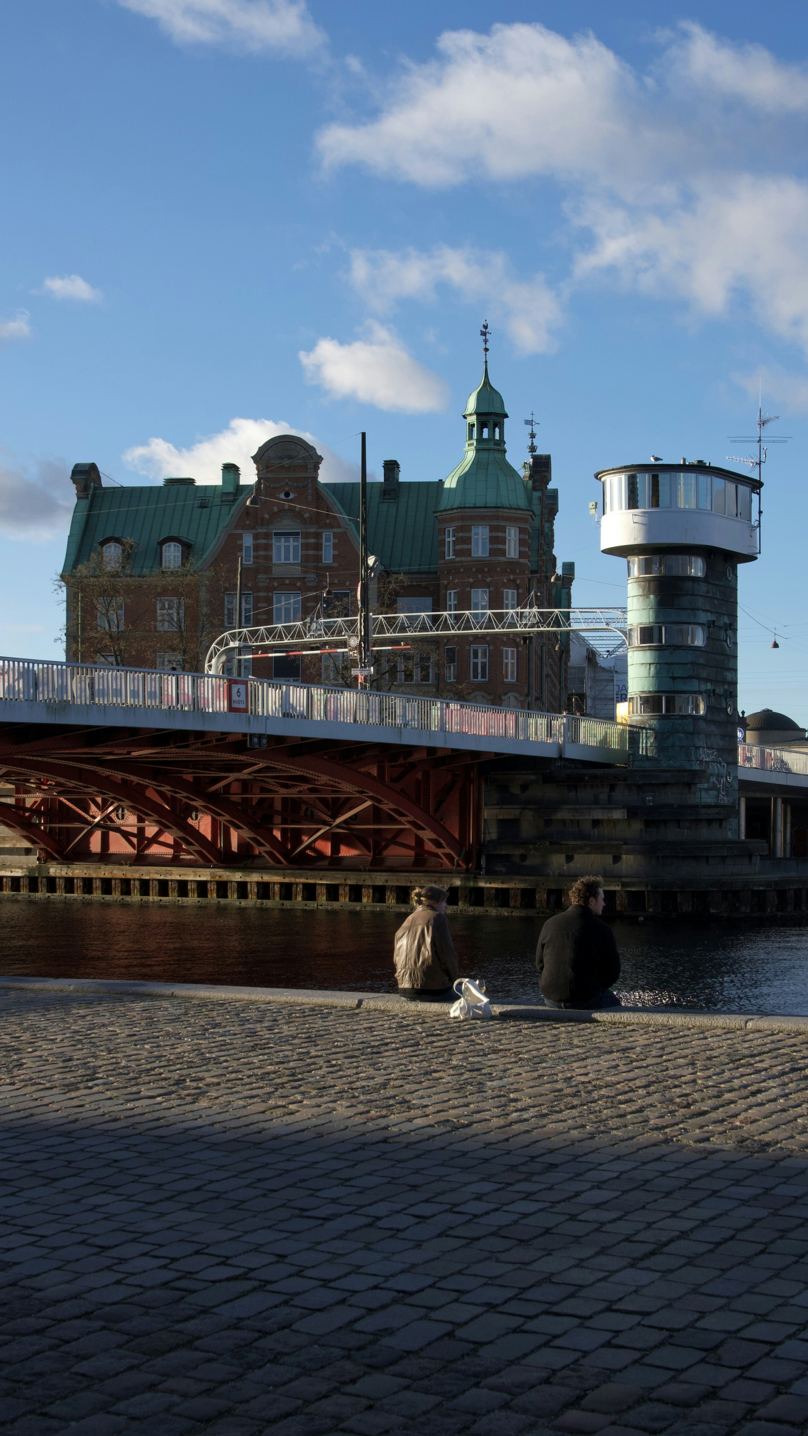 Two people sit by a waterfront with buildings and bridge