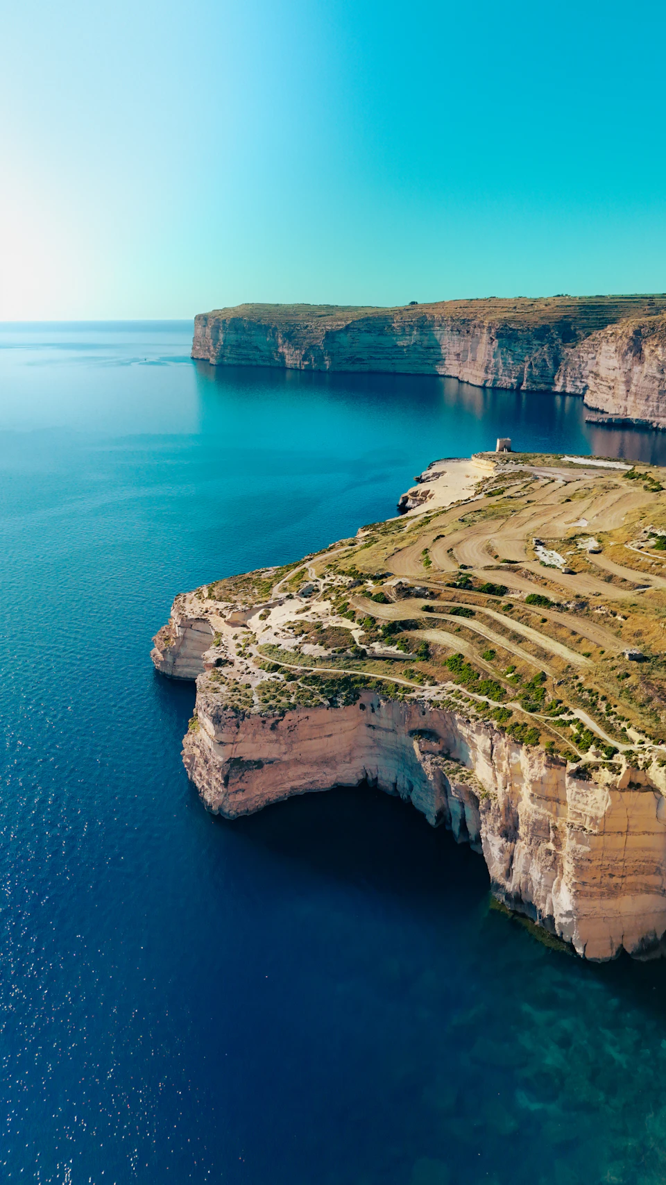 Coastal cliff with layered rock and soil formations, surrounded by deep blue-green water