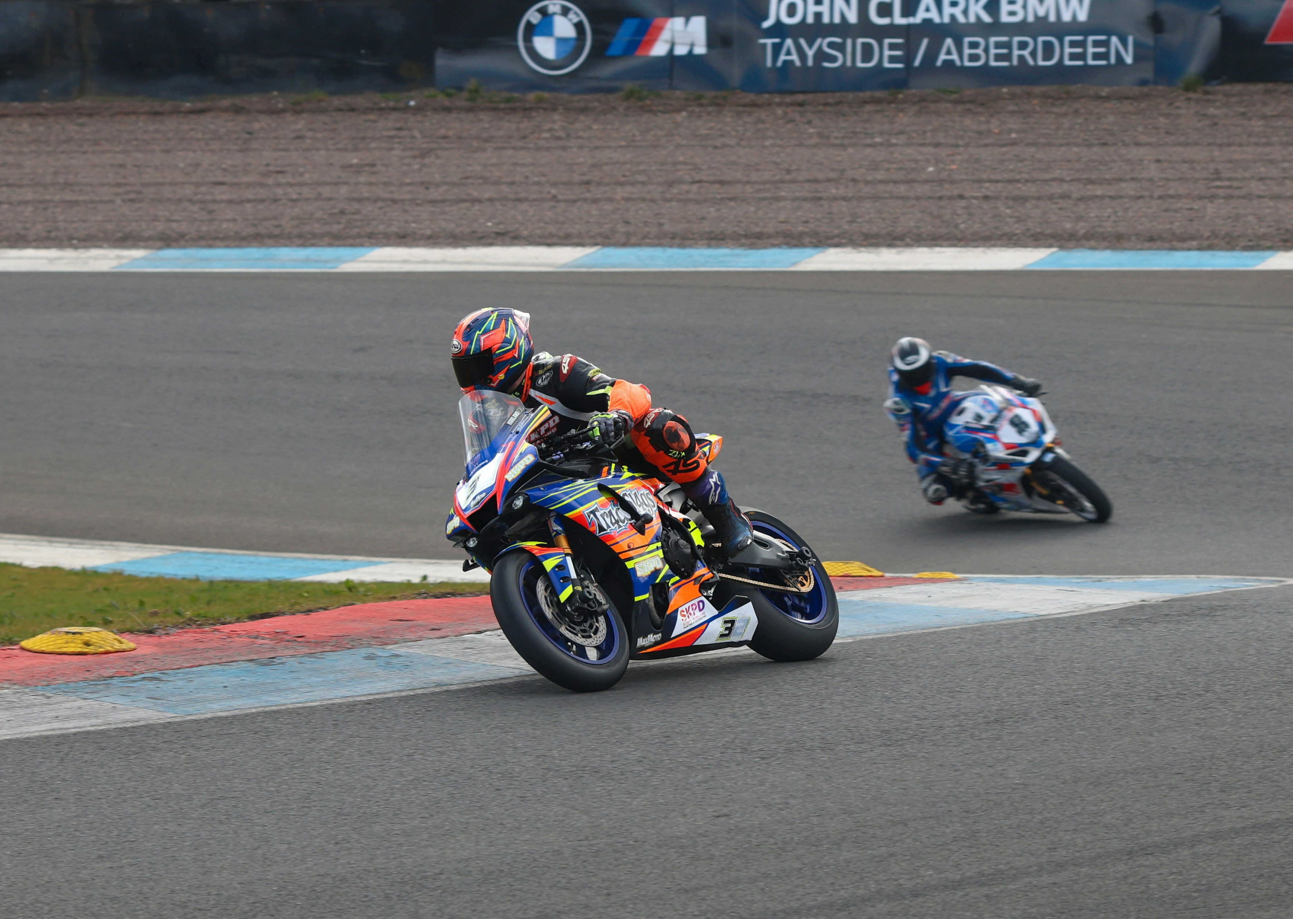 Motorcyclists race on a track with spectators in the background.