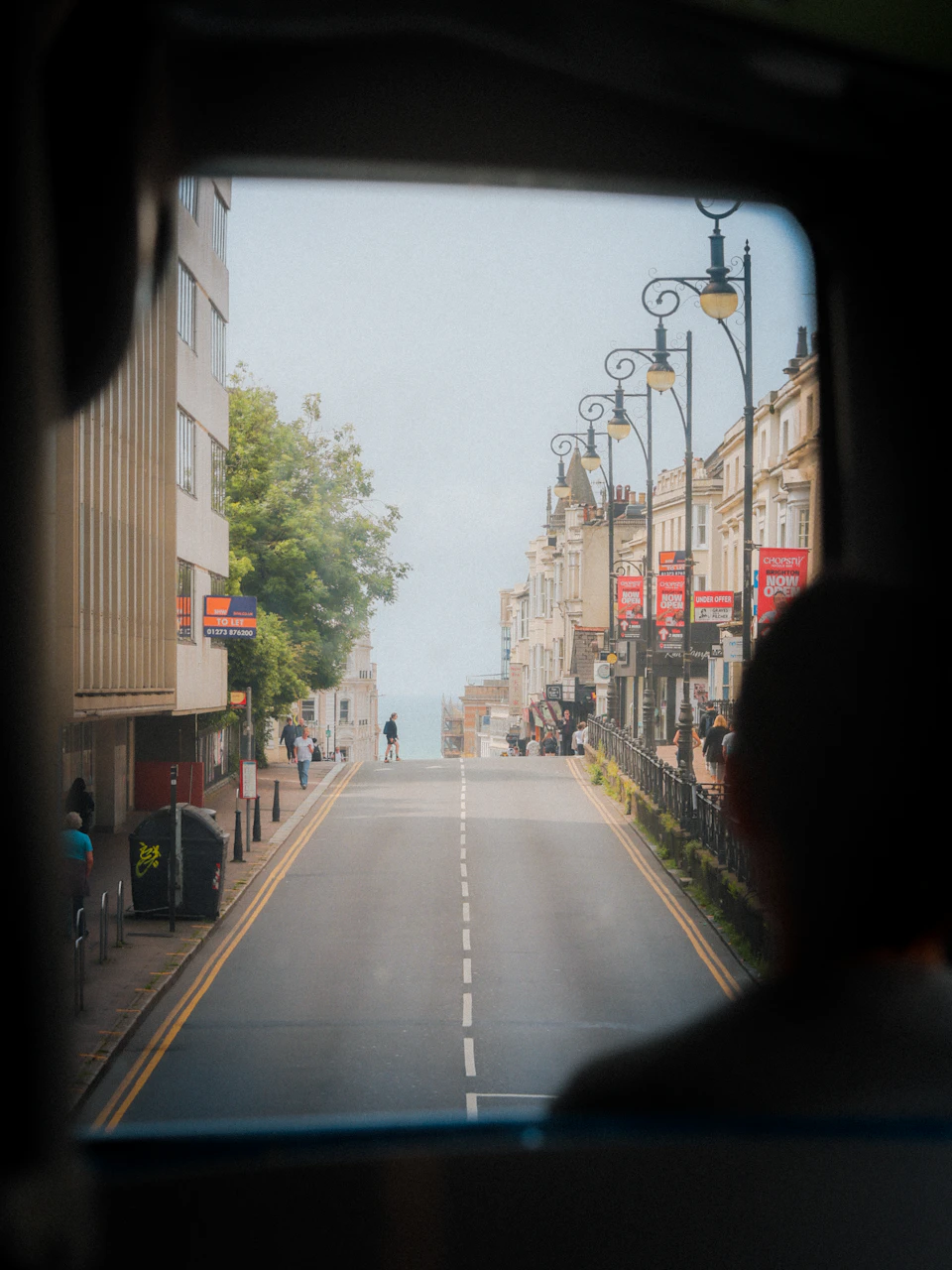 The image shows a street scene viewed through the window of a bus, with buildings lining both sides and people walking on the sidewalks. The sky is overcast, casting a soft light over the cityscape. A person can be seen standing near the center of the frame, adding depth to the scene.