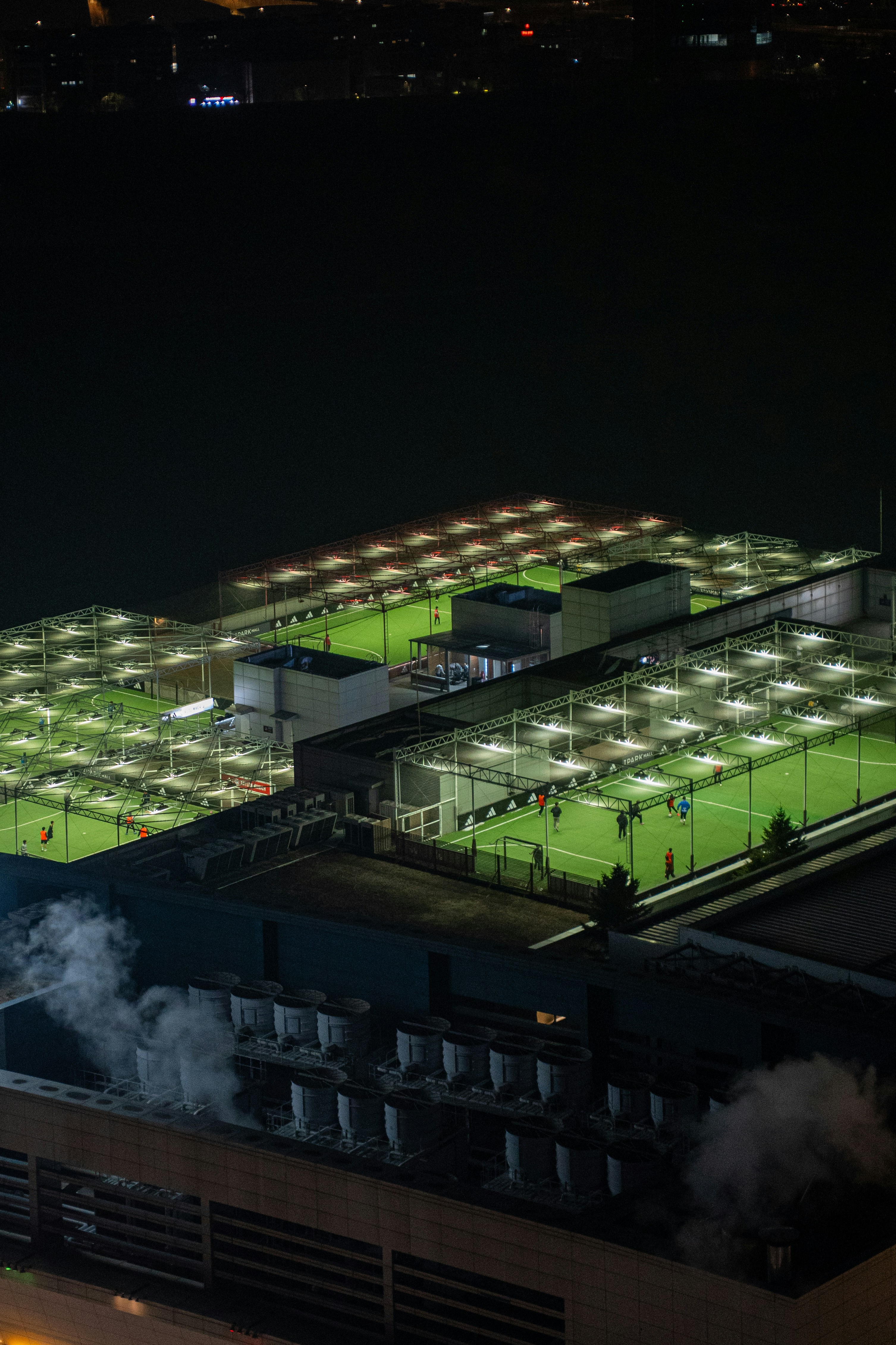 Illuminated rooftop soccer fields at night with city lights.