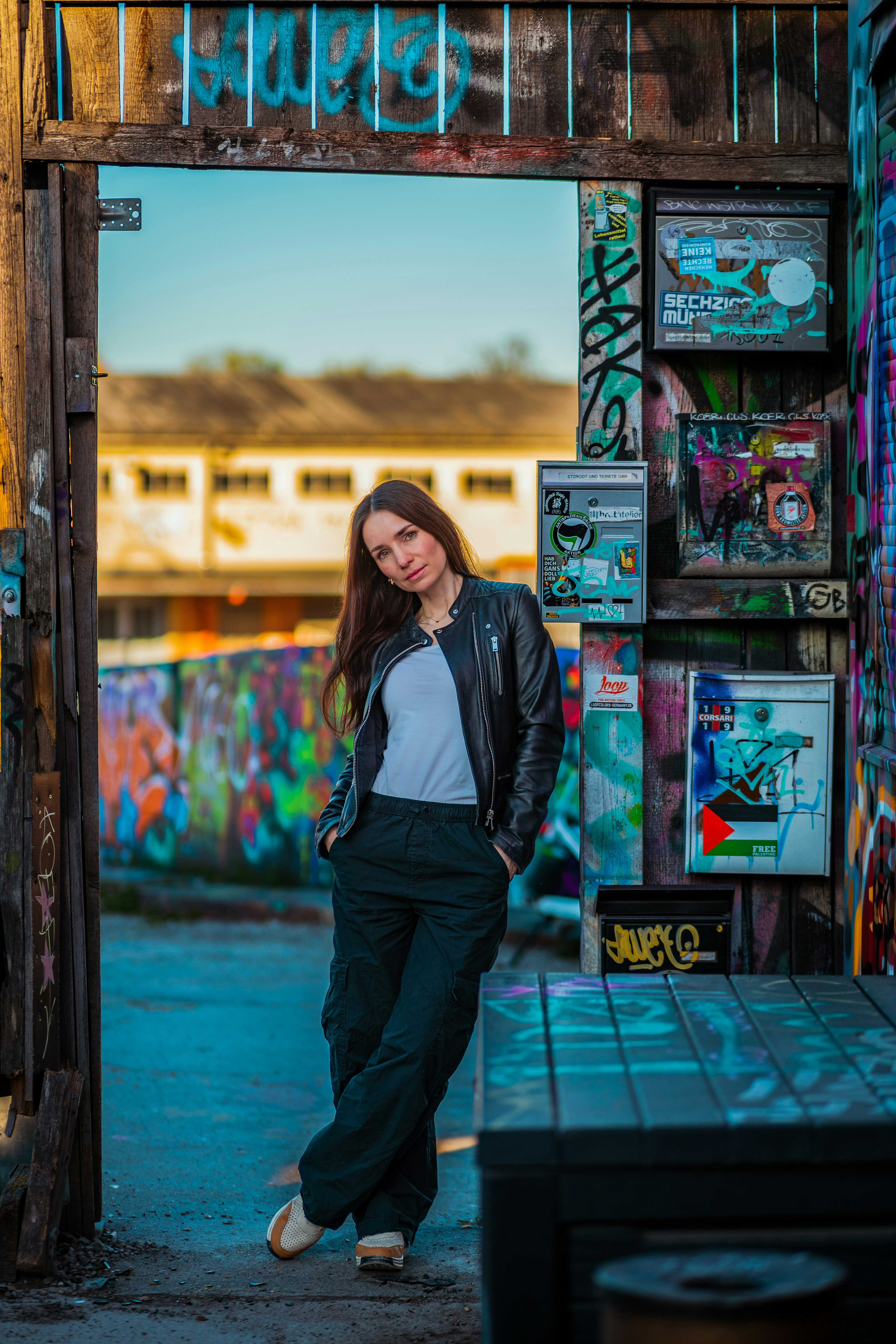 Woman in leather jacket leans against graffiti-covered wall.