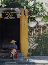 Woman in conical hat sits outside shop with lanterns.