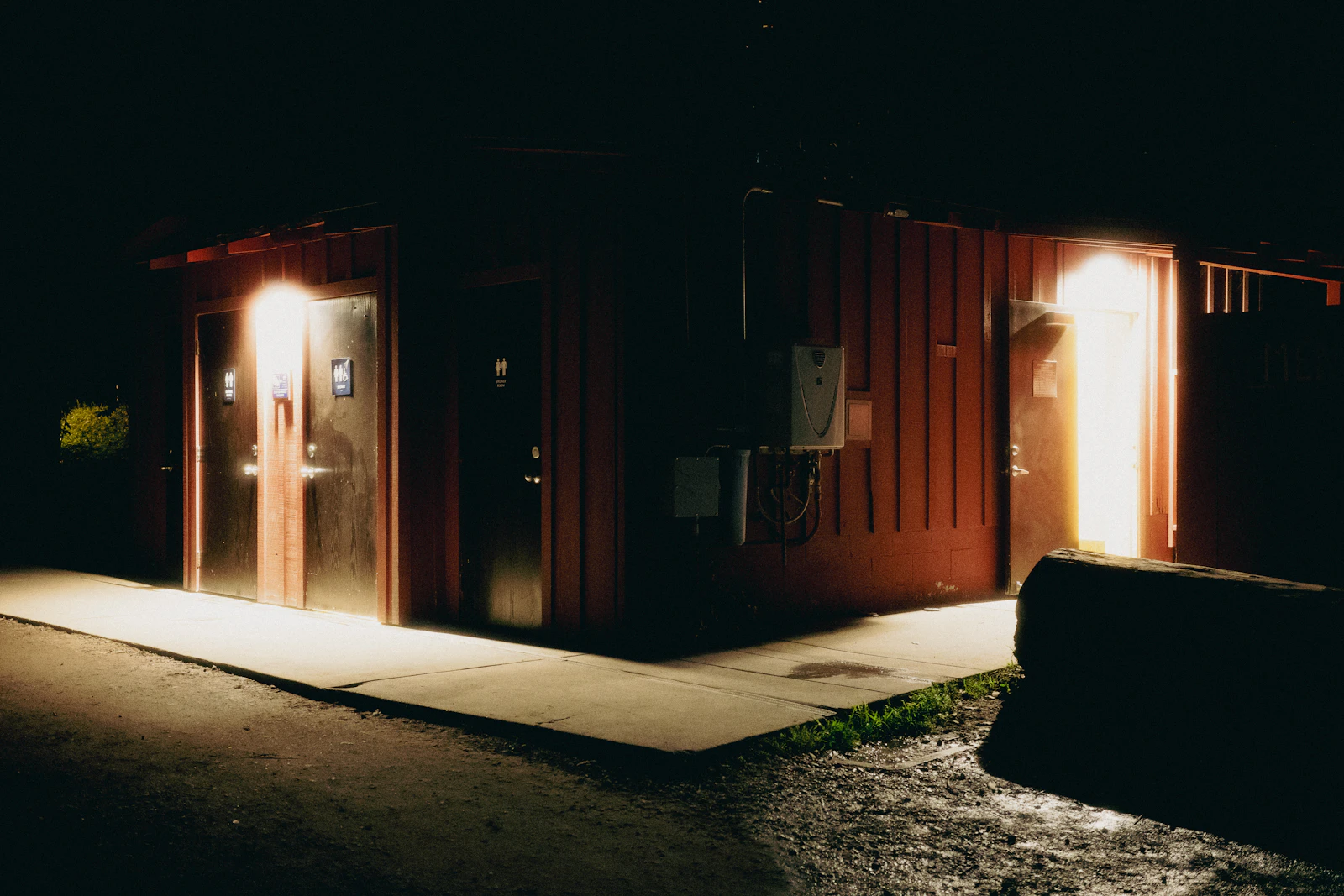 A dark red campground bathhouse lit from inside at night, with restroom doors and a concrete walkway running past the corner of the building