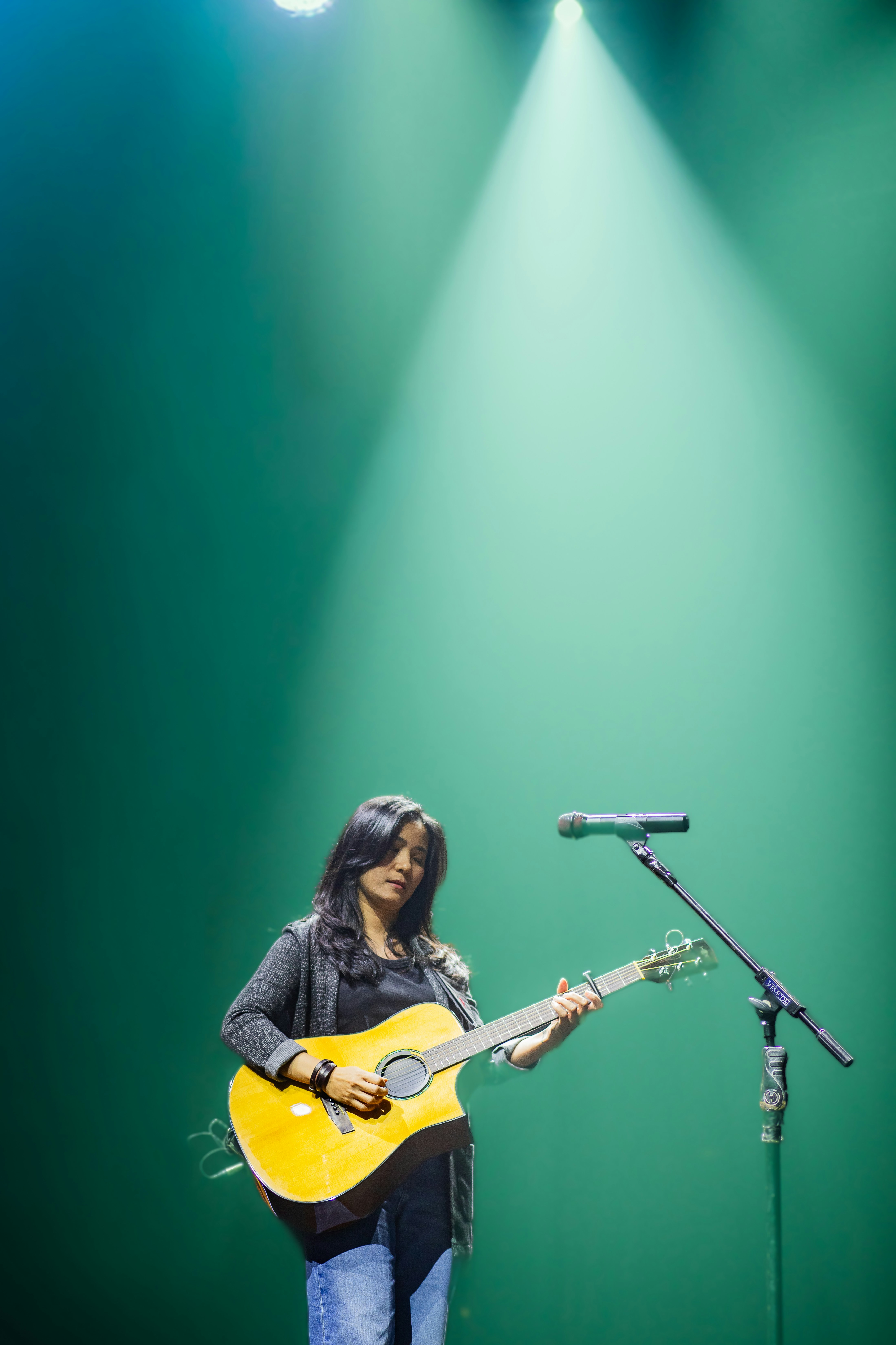 Wanita bermain gitar akustik di atas panggung di bawah sorotan.