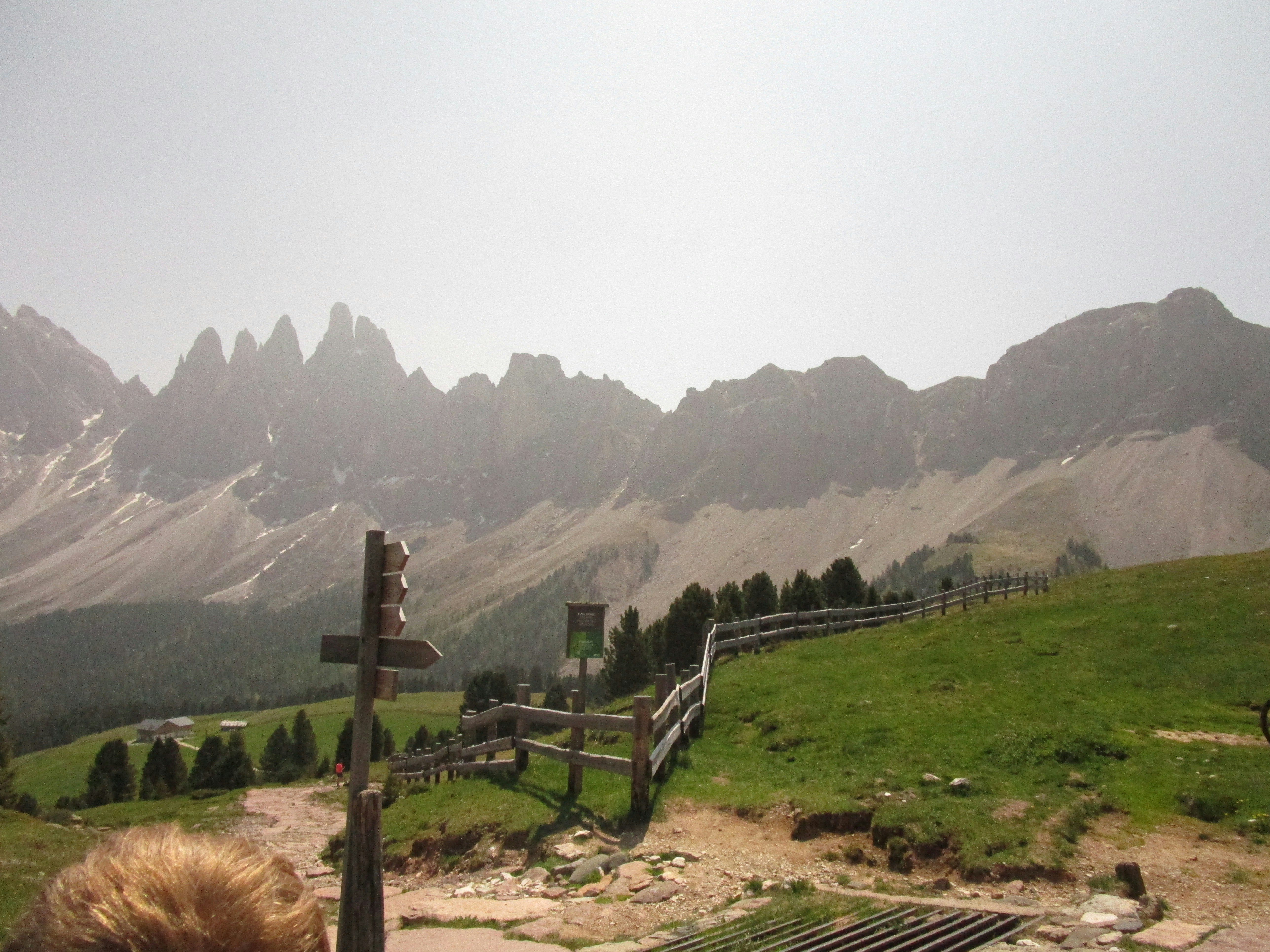 Trail winds toward jagged mountains under bright sky; foreground shows grassy path and a wooden fence guiding the eye toward distant peaks. Illustration-like natural landscape.