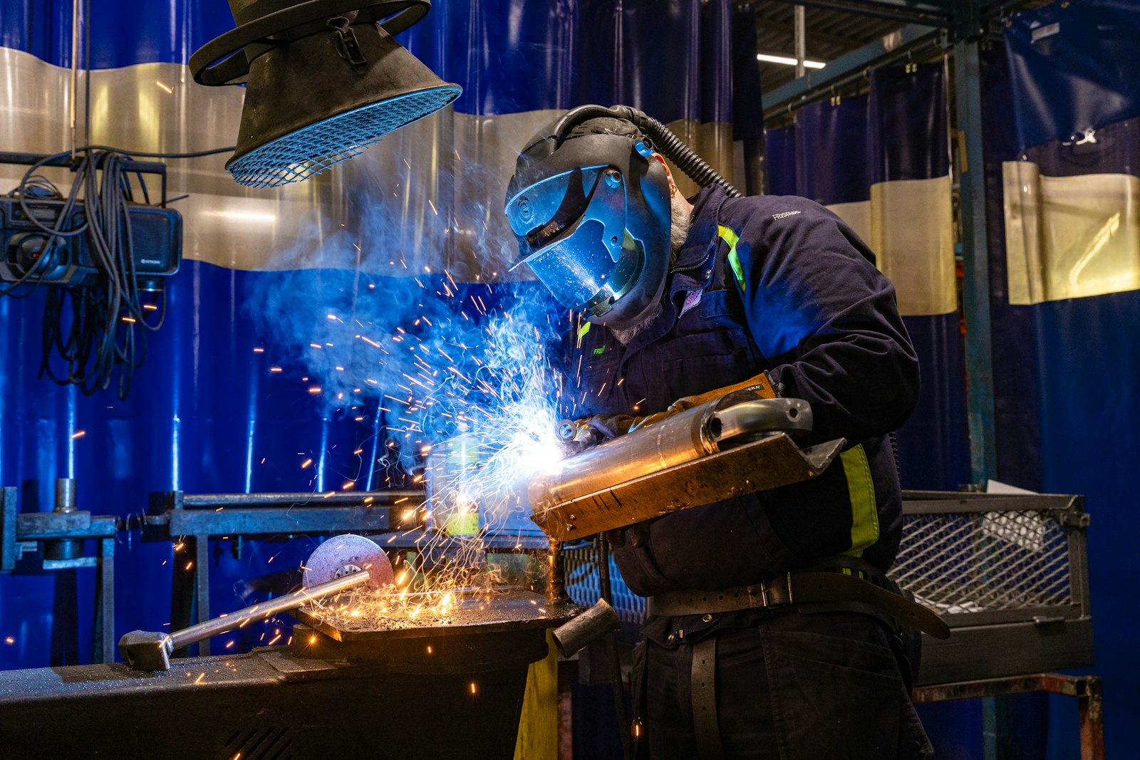 A welder in a hood, respirator and heavy gloves runs a torch along a metal part clamped to a shop bench, sparks scattering across the frame.