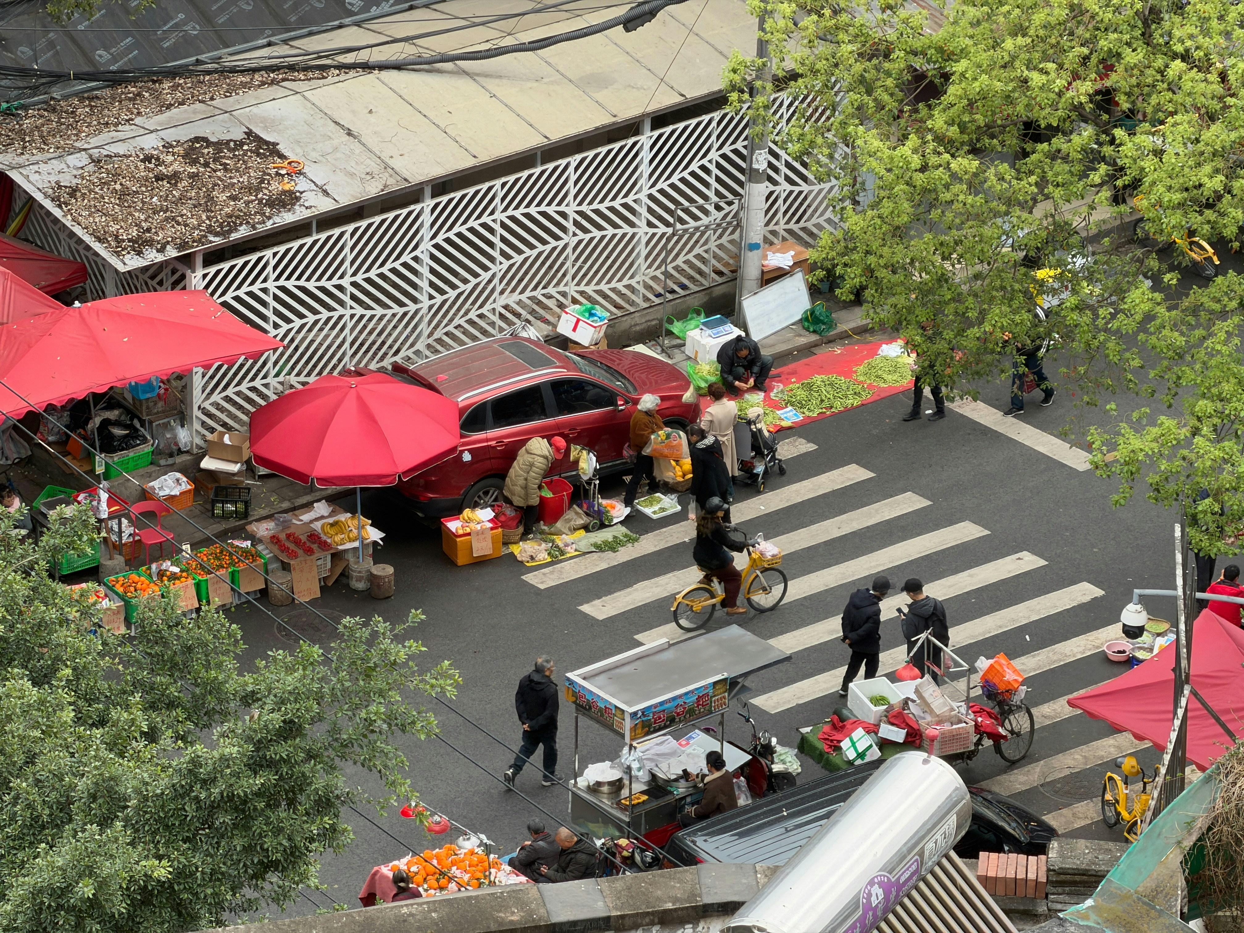 Mercado de rua com vendedores, compradores e um carro vermelho.