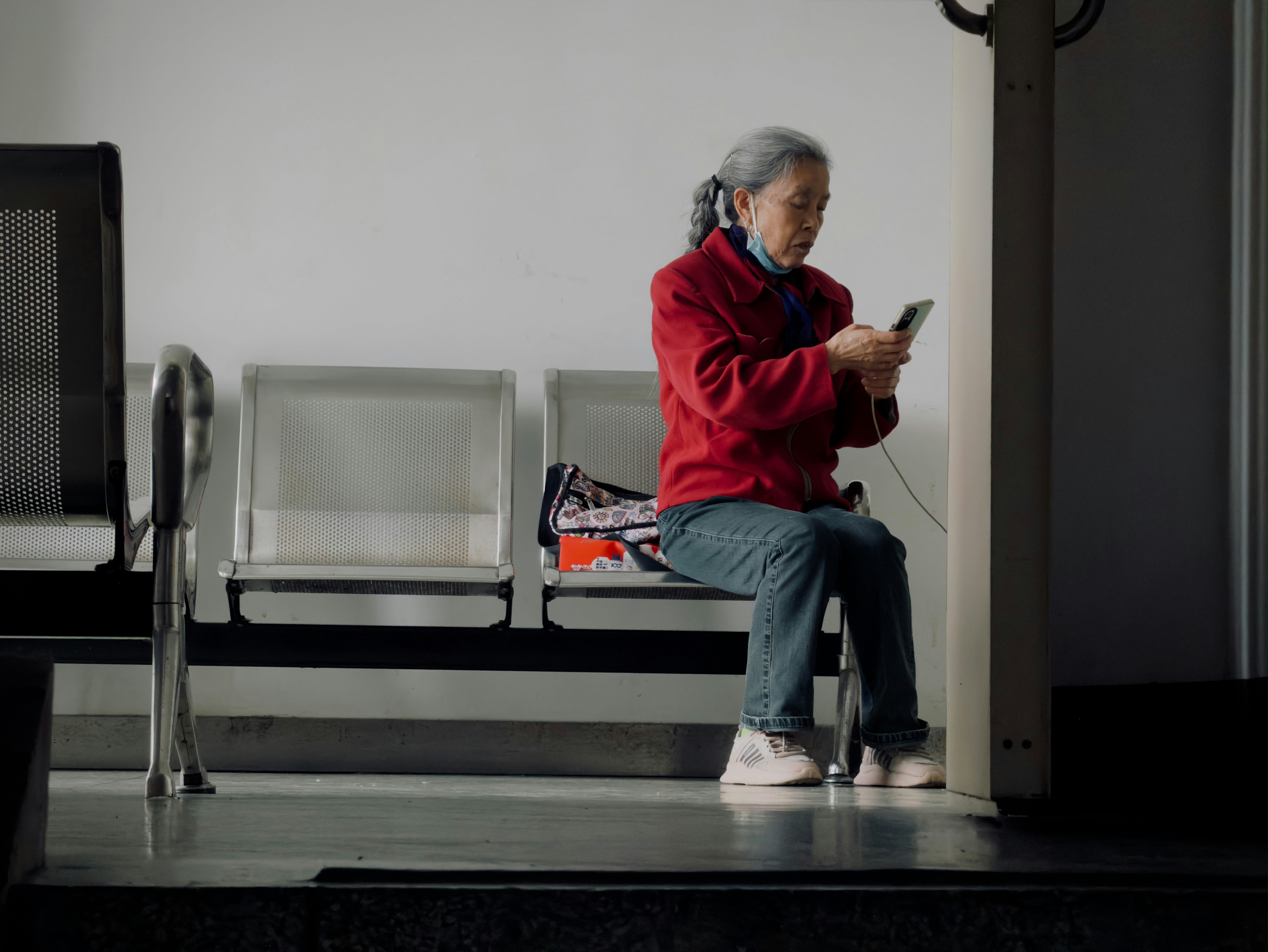 Elderly woman in red jacket uses smartphone while sitting.