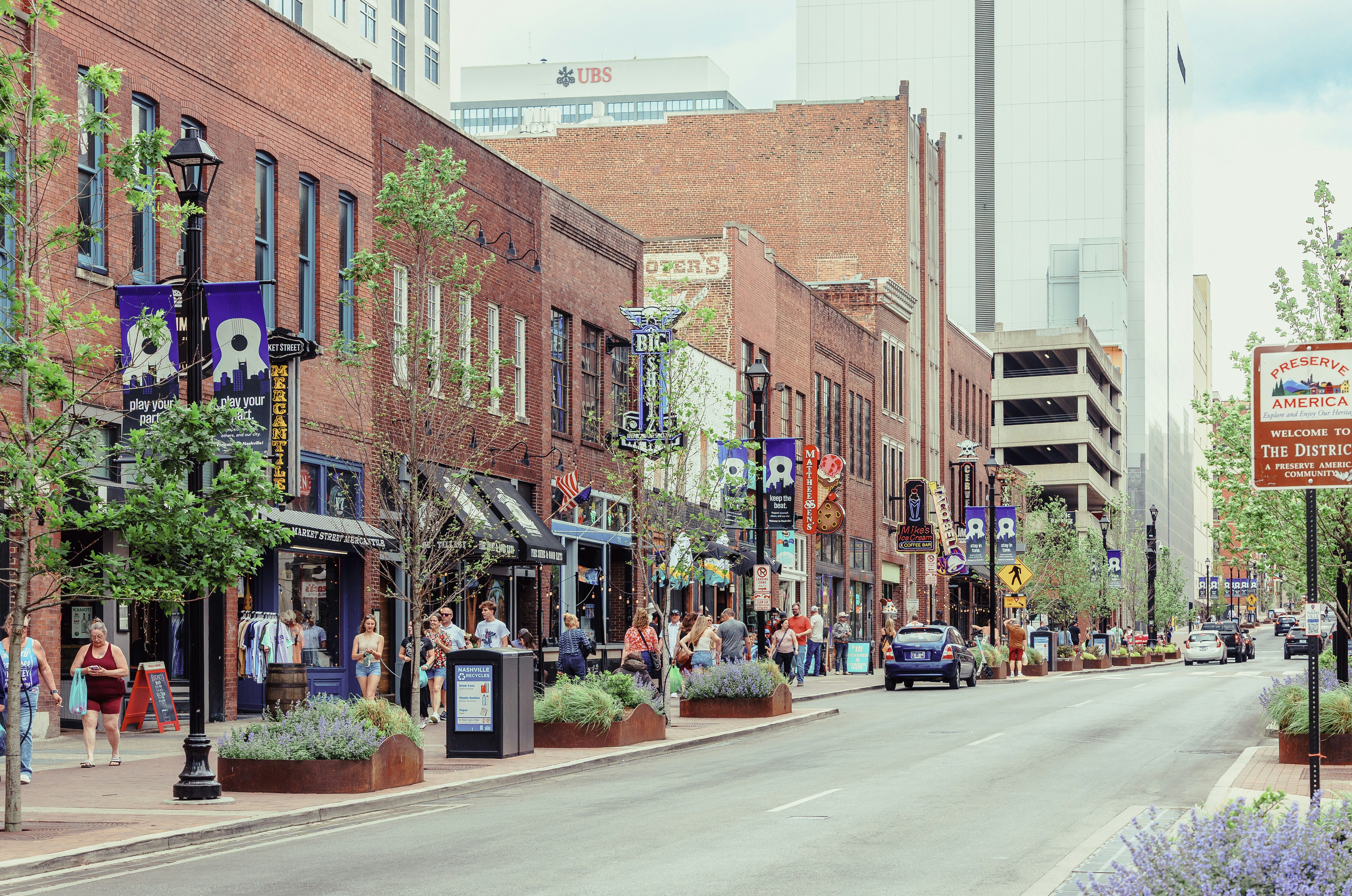 People strolling along a tree-lined street with brick buildings.