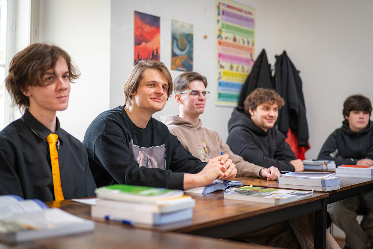 Five young men smiling along one side of a classroom table stacked with books, a colour-coded chart on the wall behind