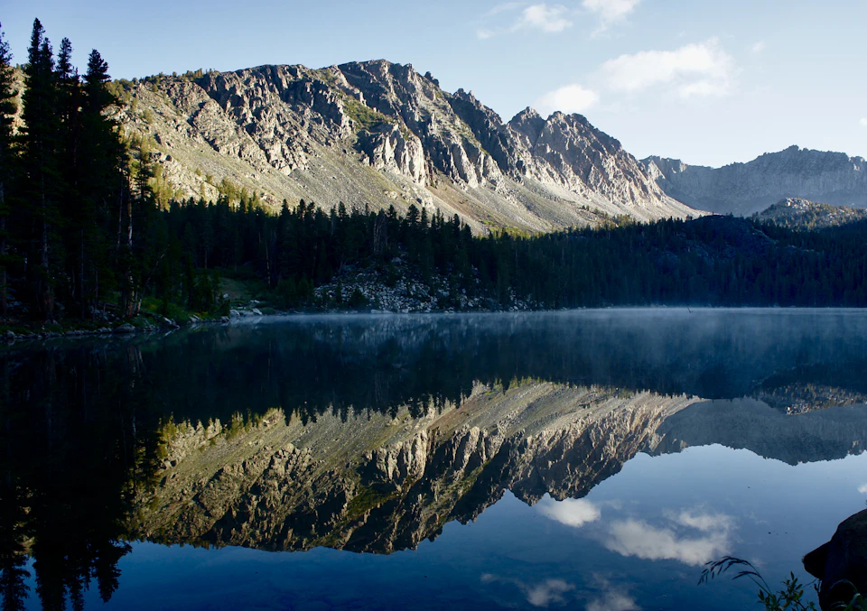 The image depicts a serene mountain lake surrounded by lush green trees and majestic mountains. The calm, light blue water reflects the surrounding landscape, creating a mirror-like effect that enhances the beauty of the scene. The mountains, with their gray and brown hues, rise majestically in the background, adding depth to the image. The sky above is a clear blue, dotted with fluffy white clouds, completing the picturesque setting.