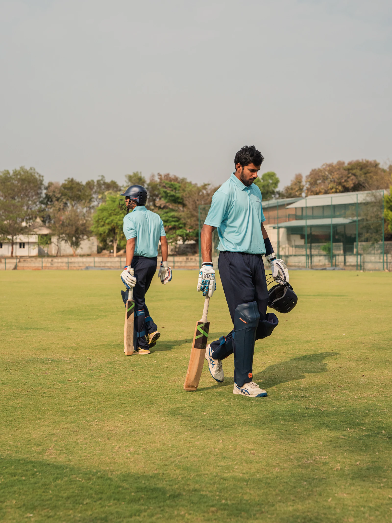 Two cricketers carrying bats walking on a sunny green field