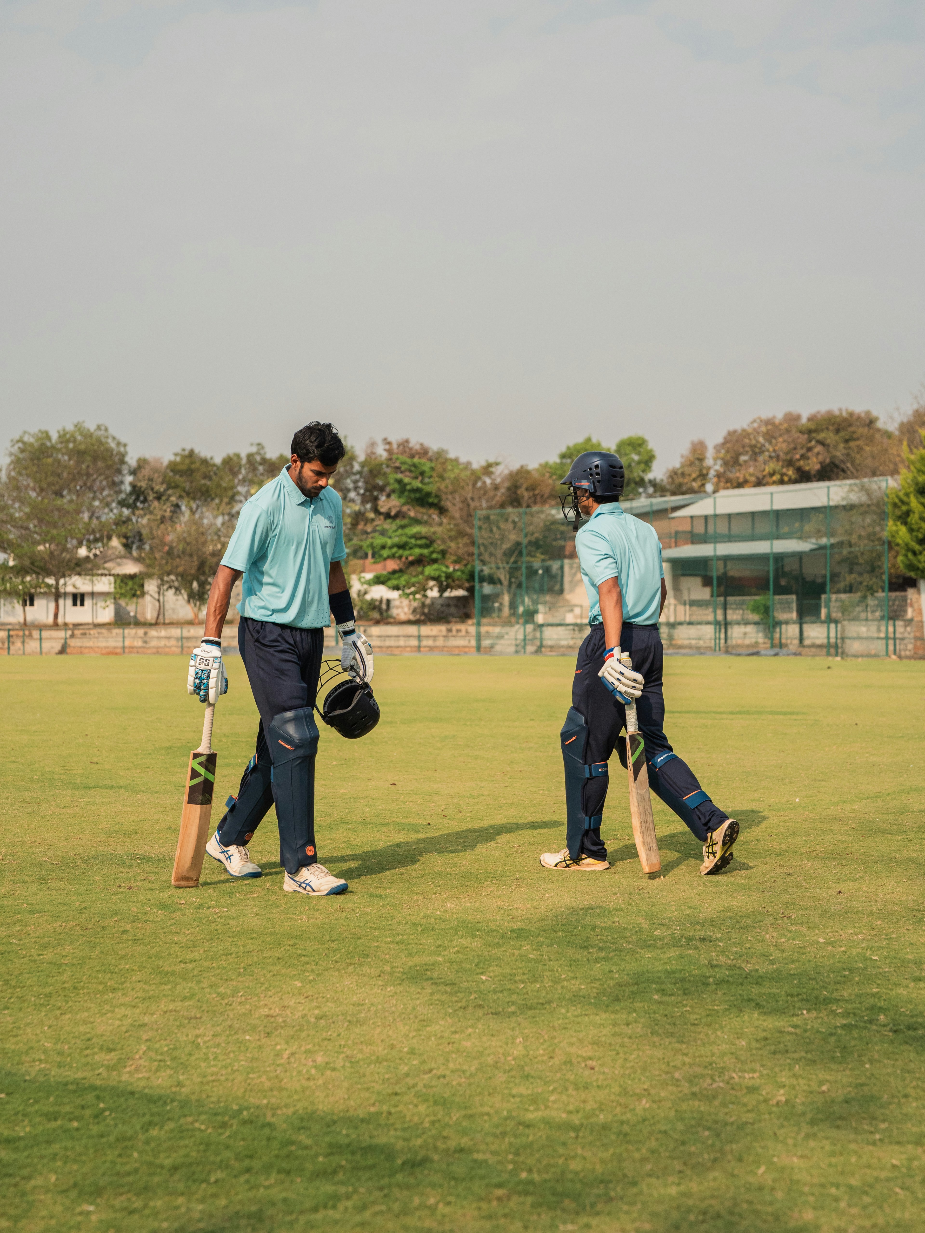 Two players in matching teal shirts walk across a sunlit cricket field, gloves in hand and bats resting at their sides, with a distant fence and trees in the background. A logo or team branding is not visible.