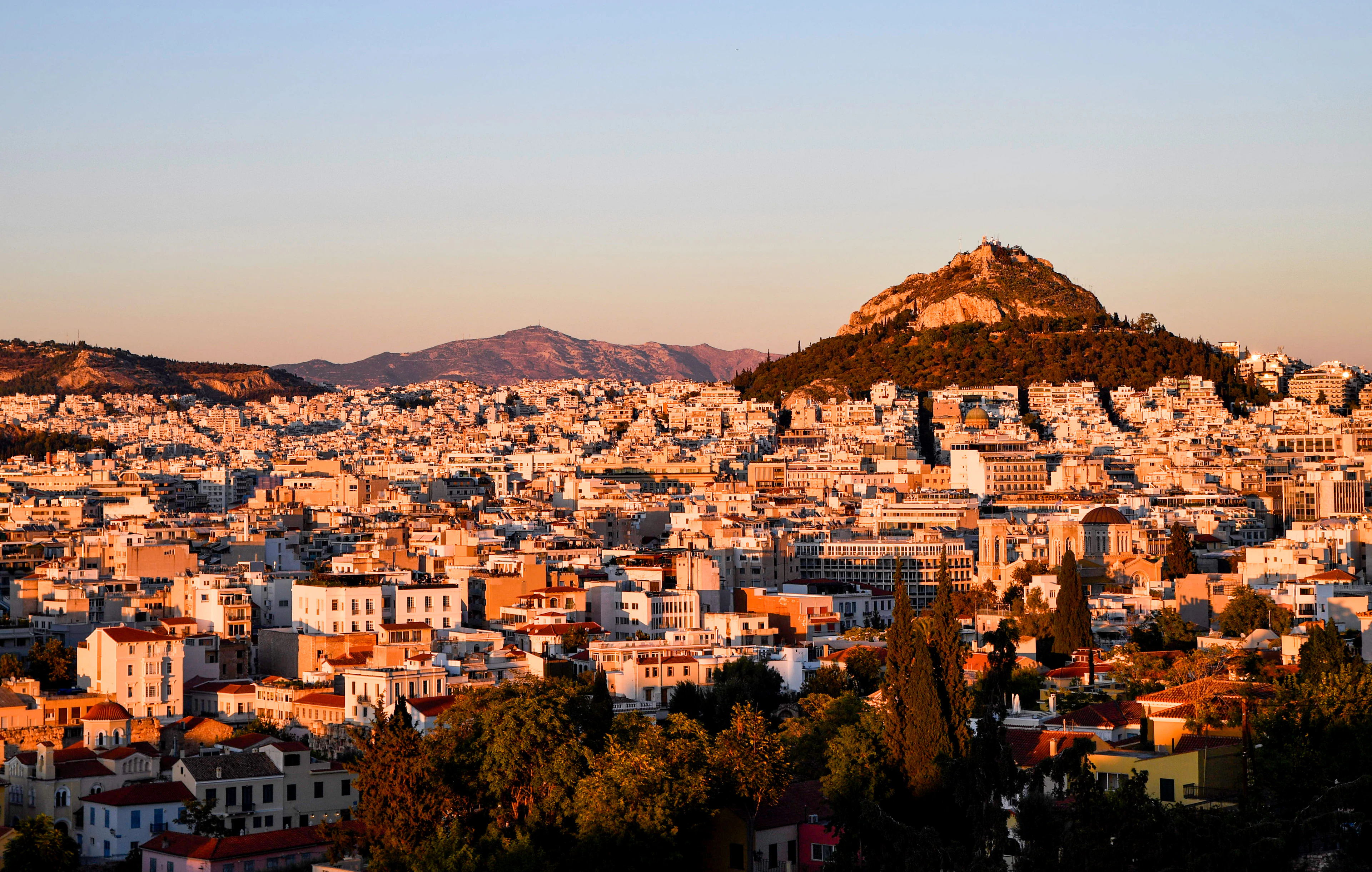 The low white rooftops of central Athens washed in golden evening light, with the pine-covered cone of Mount Lycabettus rising behind them and dry mountains on the far horizon