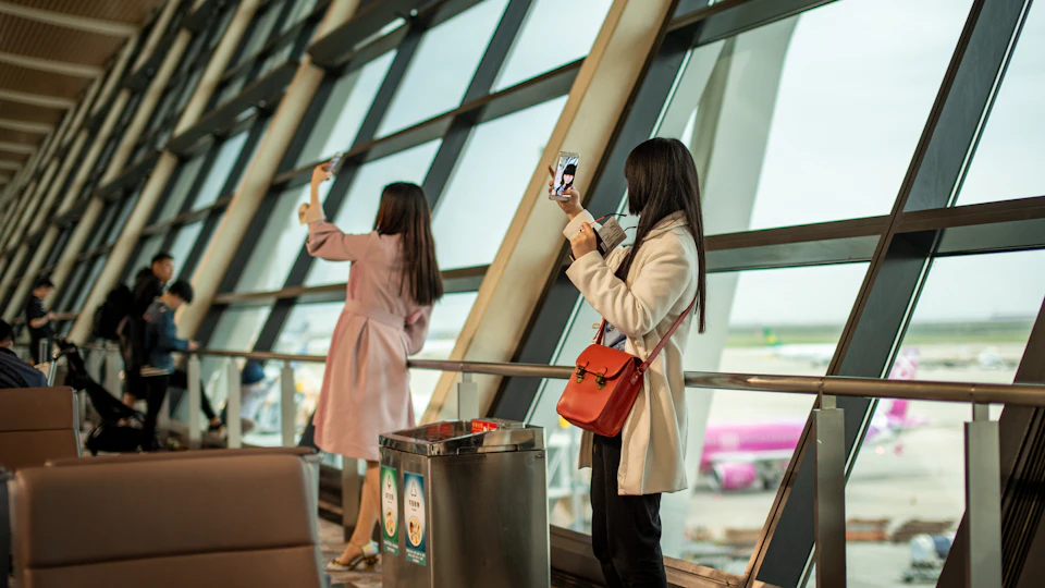 Dy udhëtare bëjnë foto me telefonat pranë një dritareje të terminalit në aeroportin Shanghai Pudong, me avionë të parkuar jashtë.
