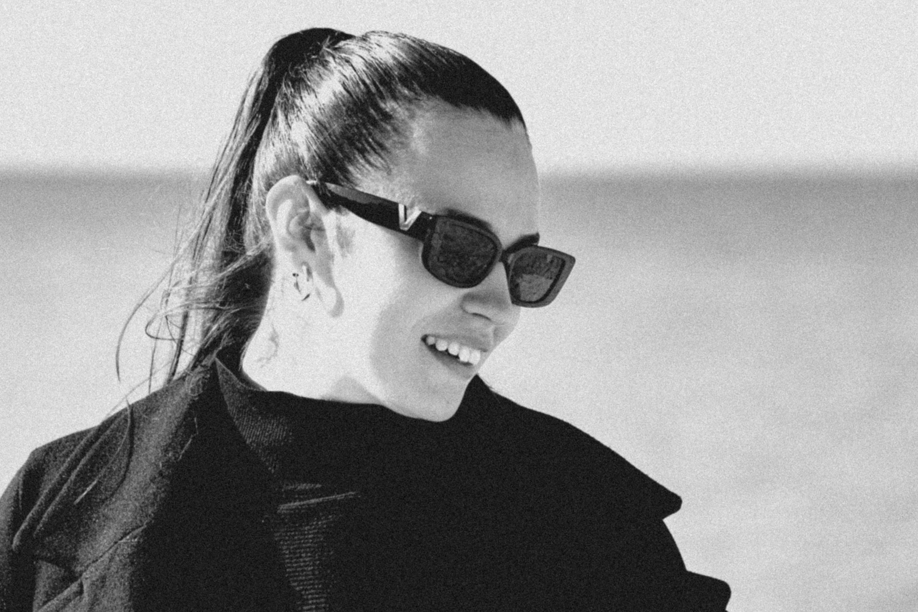 Wanita dengan kacamata hitam dan kuncir kuda tersenyum di luar ruangan