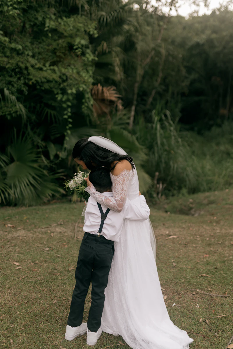 A bride in her wedding dress hugging a young boy in a garden