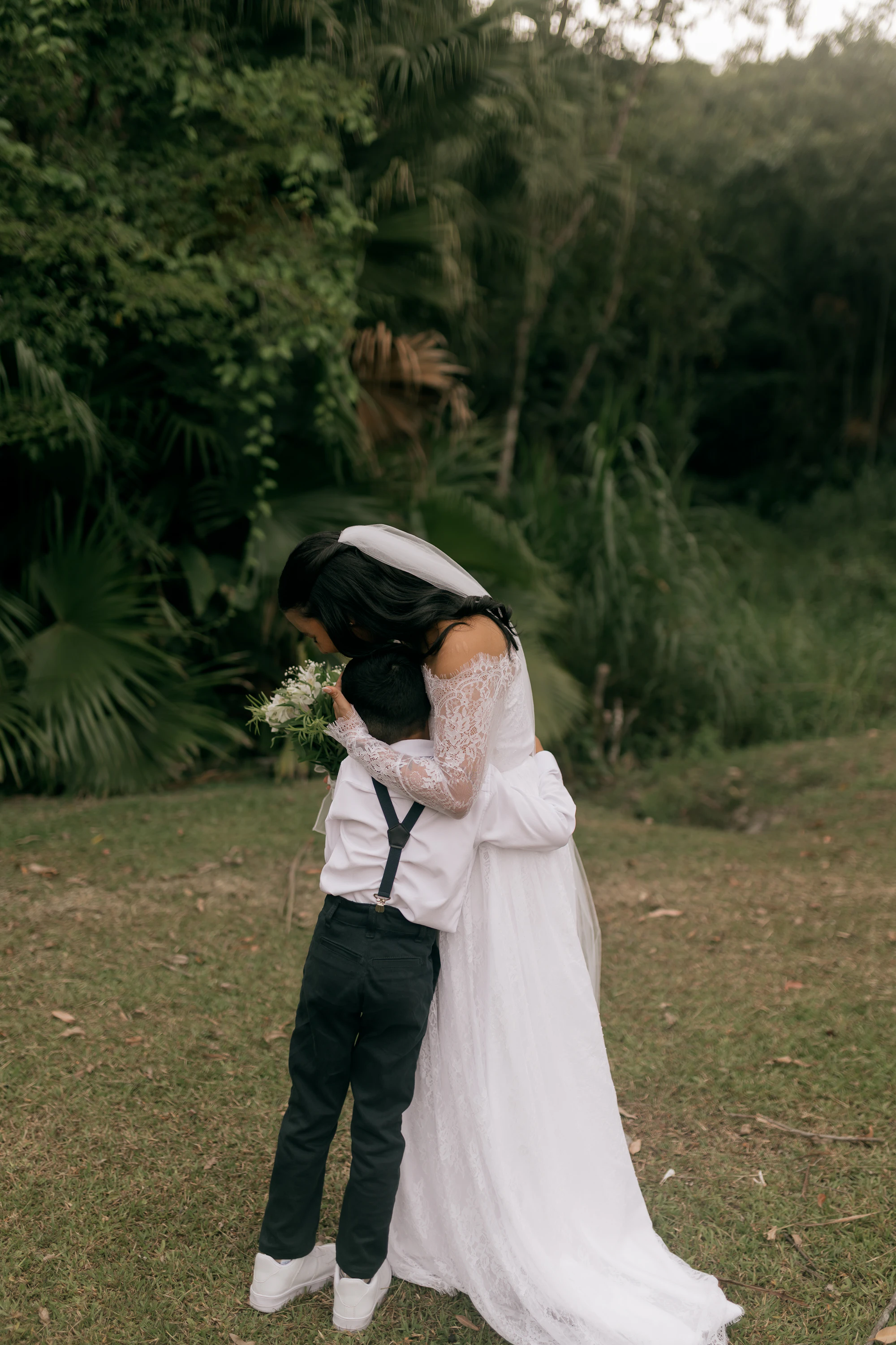 A bride in her wedding dress hugging a young boy in a garden