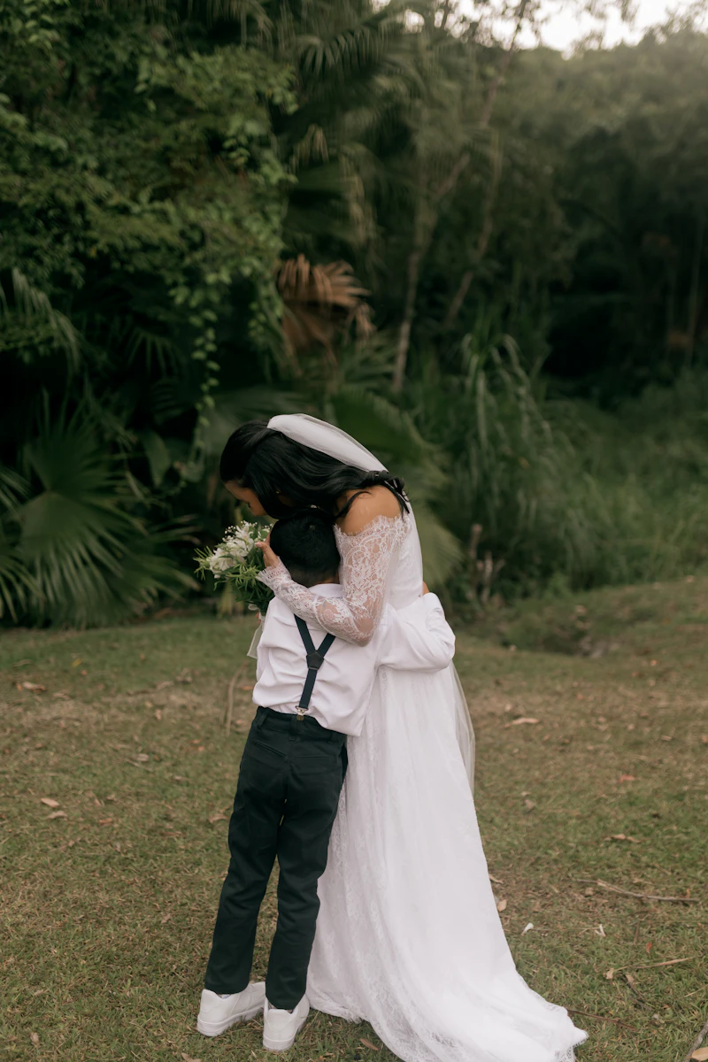 A bride in her wedding dress hugging a young boy in a garden