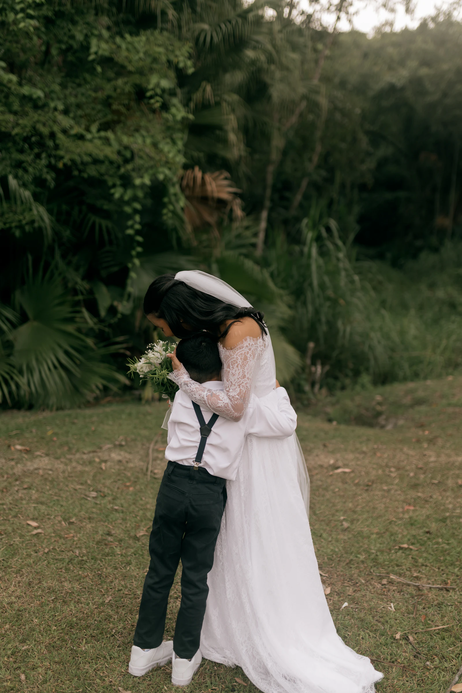 A bride in her wedding dress hugging a young boy in a garden
