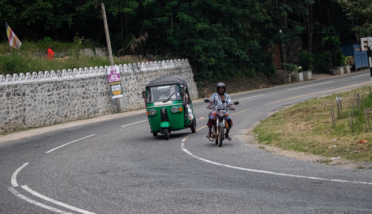 A green tuk-tuk and a motorcyclist sharing a winding road through lush Sri Lankan hill country - typical self-drive traffic on rural roads