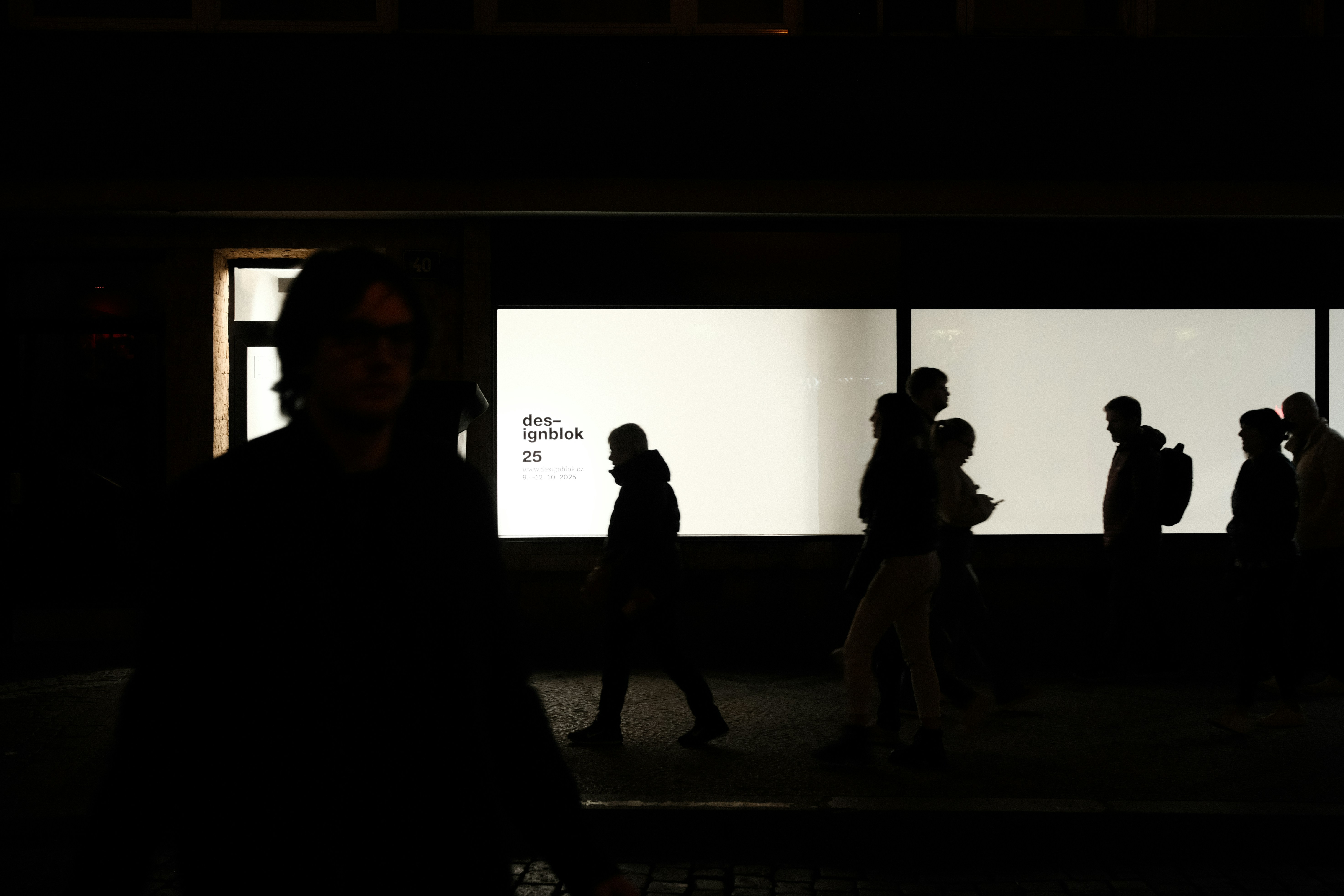Silhouettes of people walking past illuminated storefronts at night.