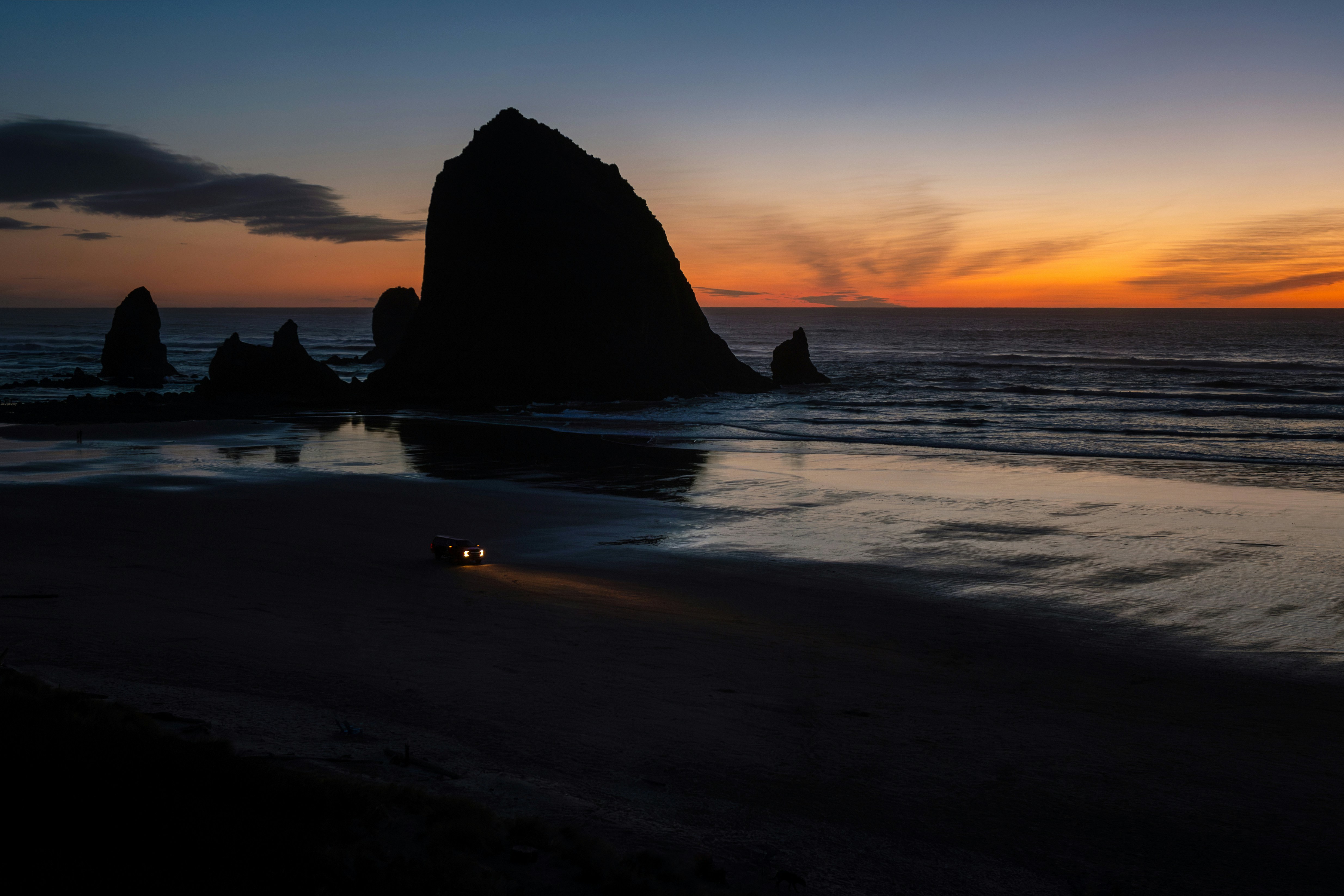 A lone car drives on a beach at dusk.