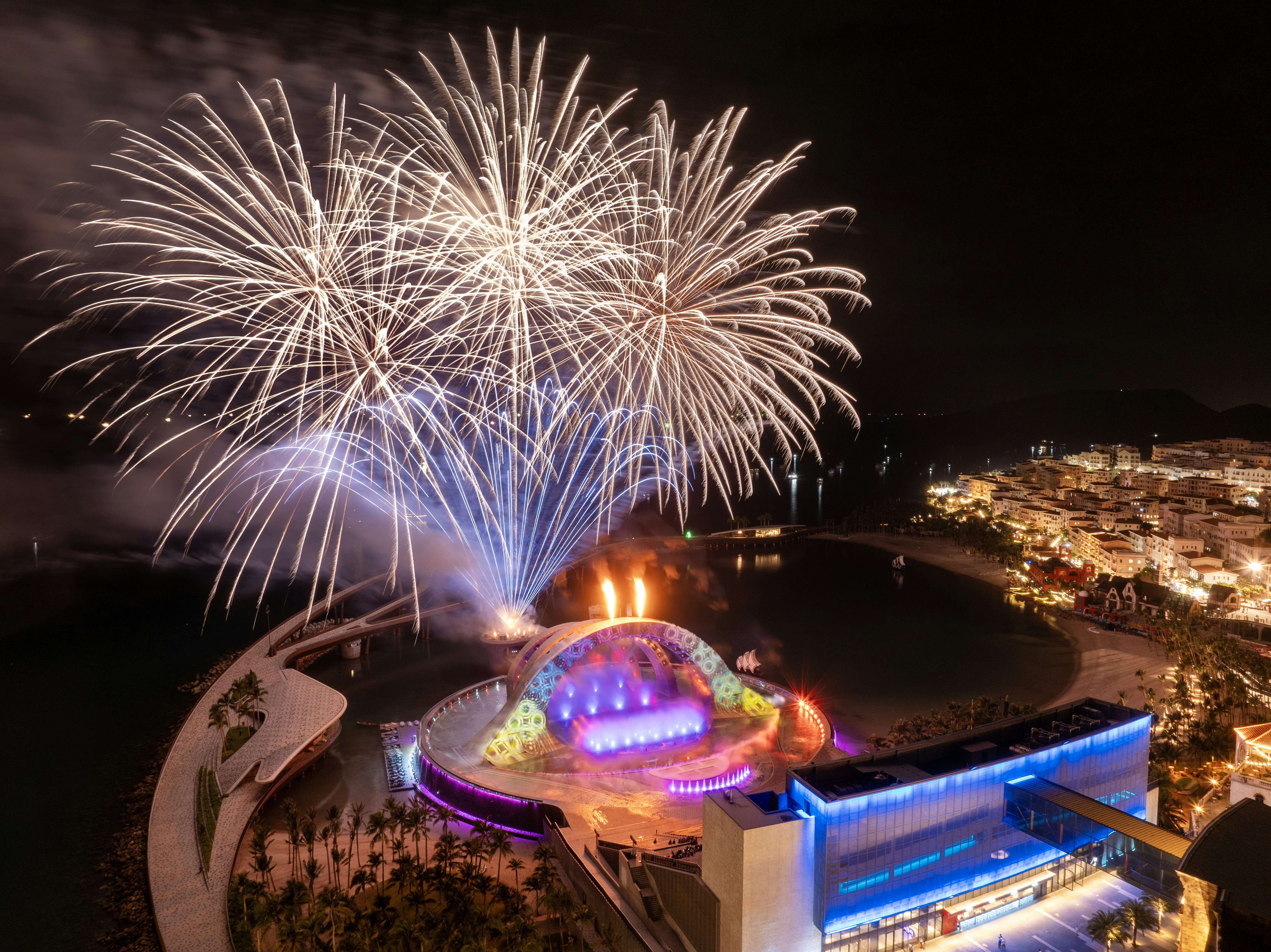Fogos de artifício explodem sobre um complexo moderno iluminado à noite.