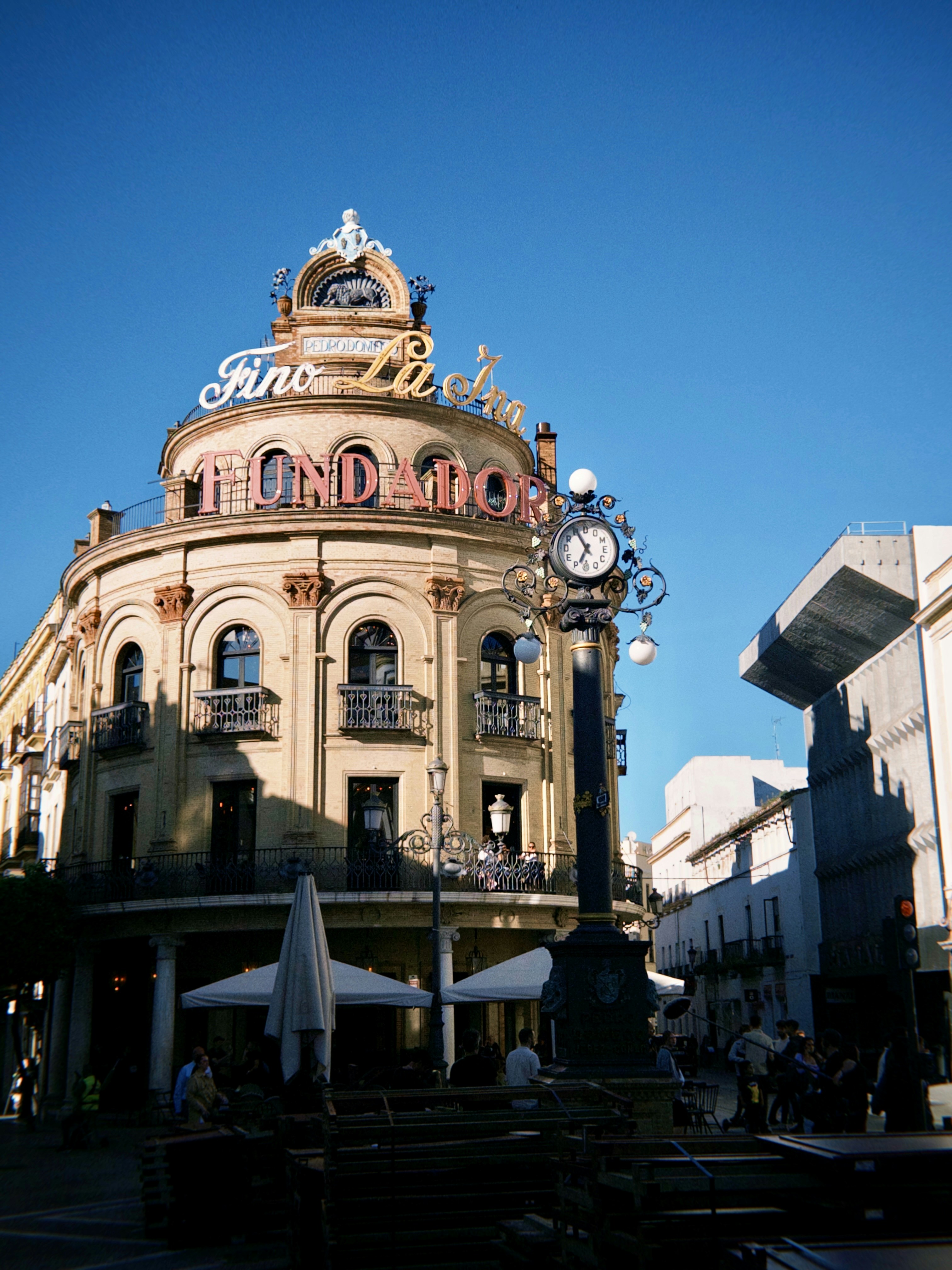The iconic El Fundador building at dusk, its terracotta lettering and neon Fino La Ina sign catching the last of the Andalusian sun. An ornate cast-iron street clock anchors the foreground, as locals and visitors fill the pavement terrace below - a scene that hasn’t changed much in a century.
