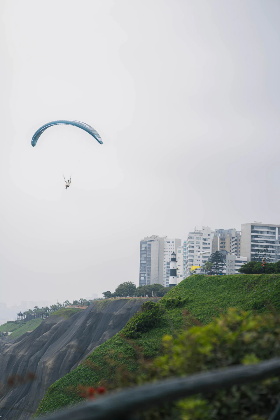 The image depicts a person paragliding over a cityscape, with a large building and several other buildings visible below them. The paraglider is soaring high above the city, performing an impressive aerial maneuver. The sky overhead is filled with clouds, creating a dramatic backdrop for this thrilling activity.