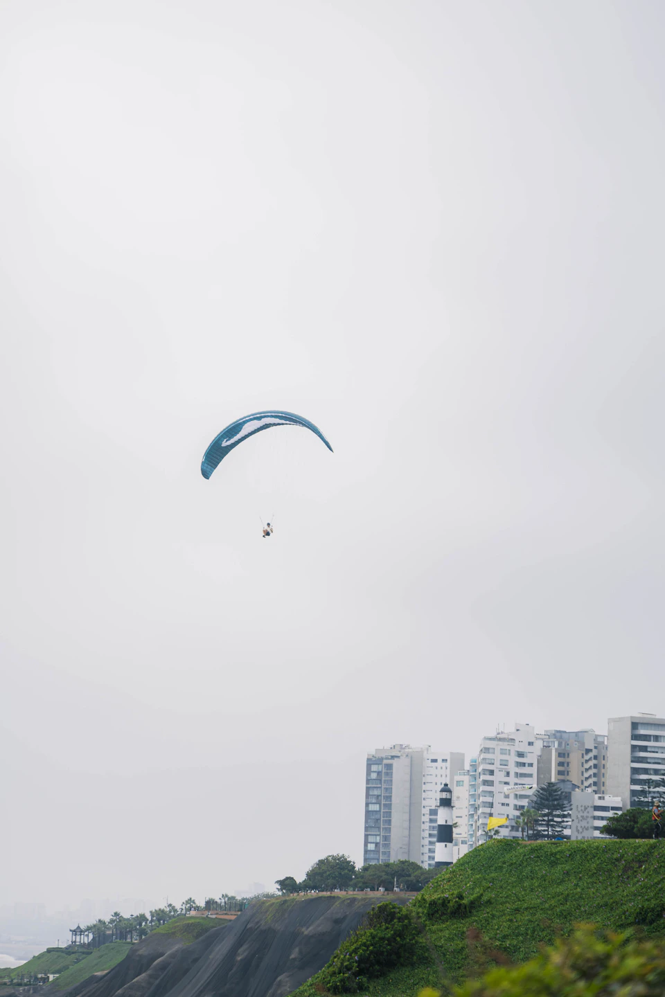 The image depicts a person paragliding over a cityscape, with a large body of water visible below them. The paraglider is soaring high above the city, performing an impressive aerial maneuver against the backdrop of the urban environment. The sky overhead is filled with clouds, creating a dramatic and moody atmosphere for this thrilling activity.

The city itself is densely populated, with numerous buildings of varying heights lining the streets below. A lighthouse can be seen in the distance, adding to the coastal setting.