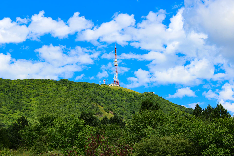 Large radio tower atop a hill, surrounded by lush greenery and fluffy white clouds