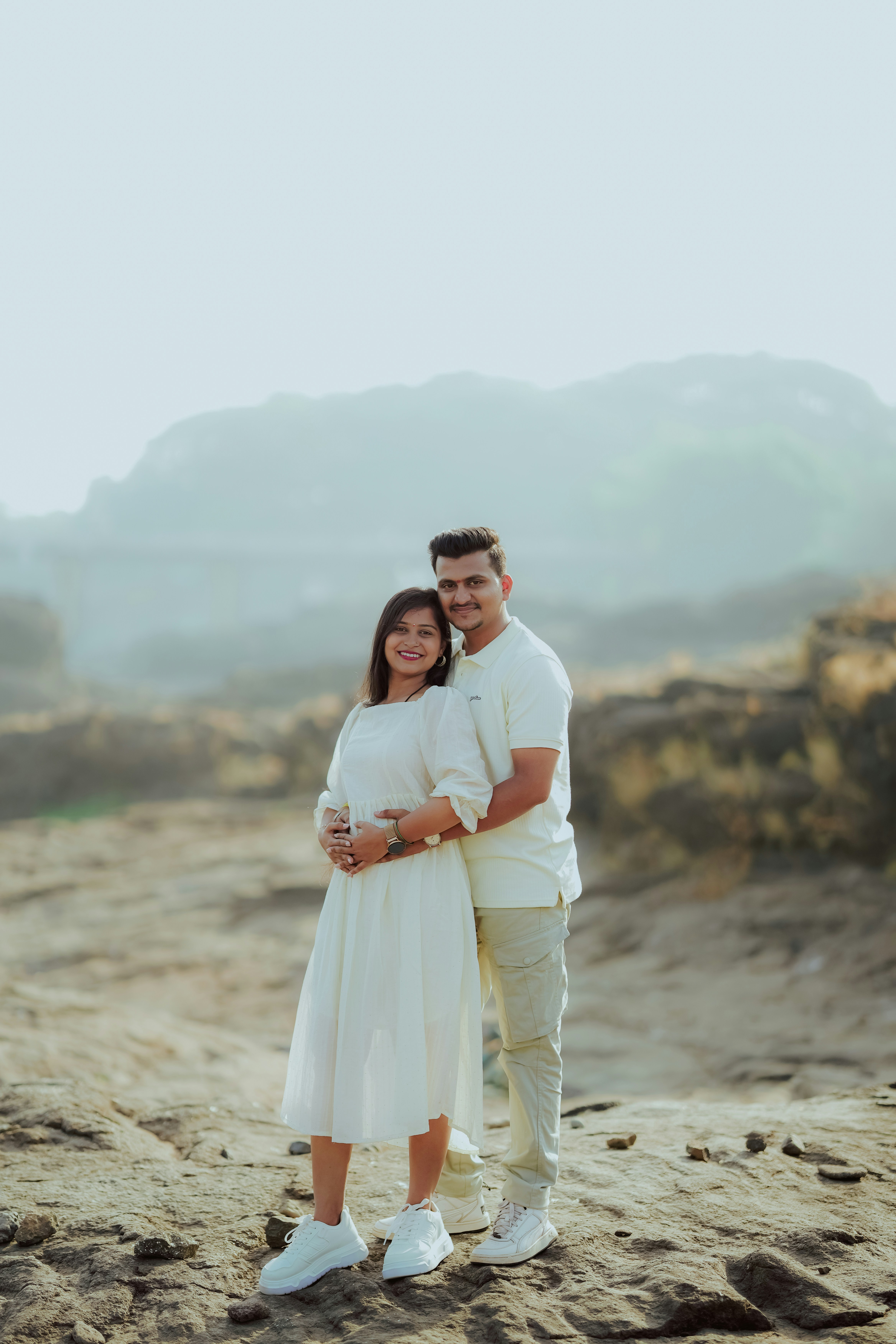 A couple embraces on a rocky terrain with mountains behind.