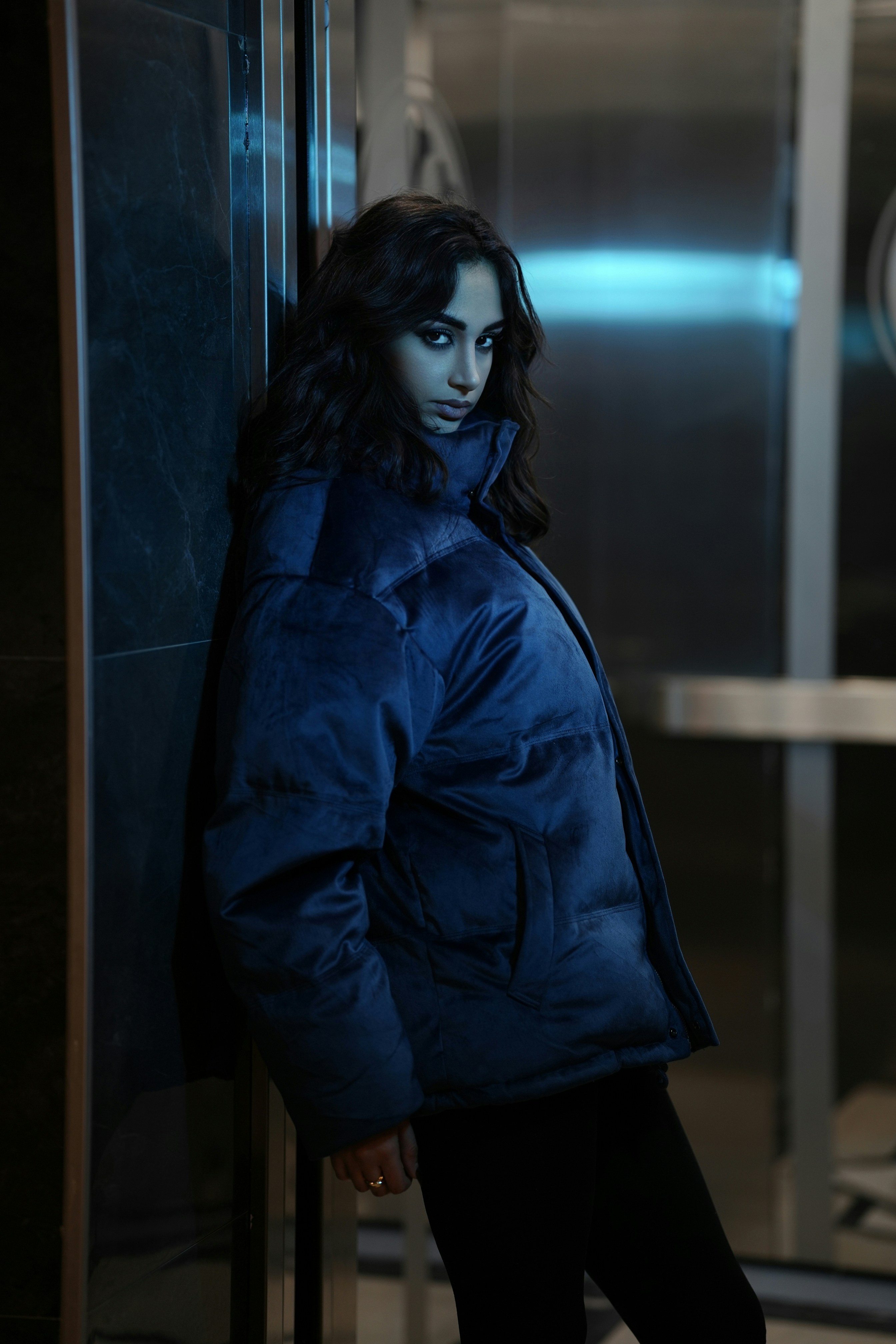 Woman in a blue puffer jacket leans against wall.