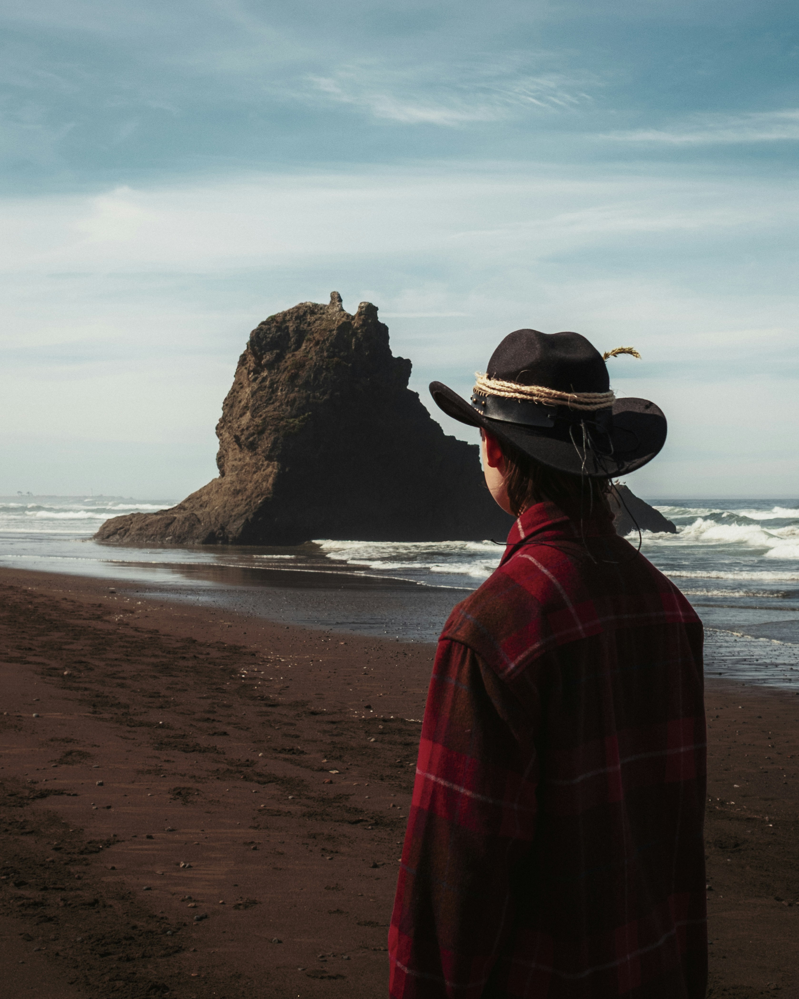 Person in hat looks at rock formation on beach.