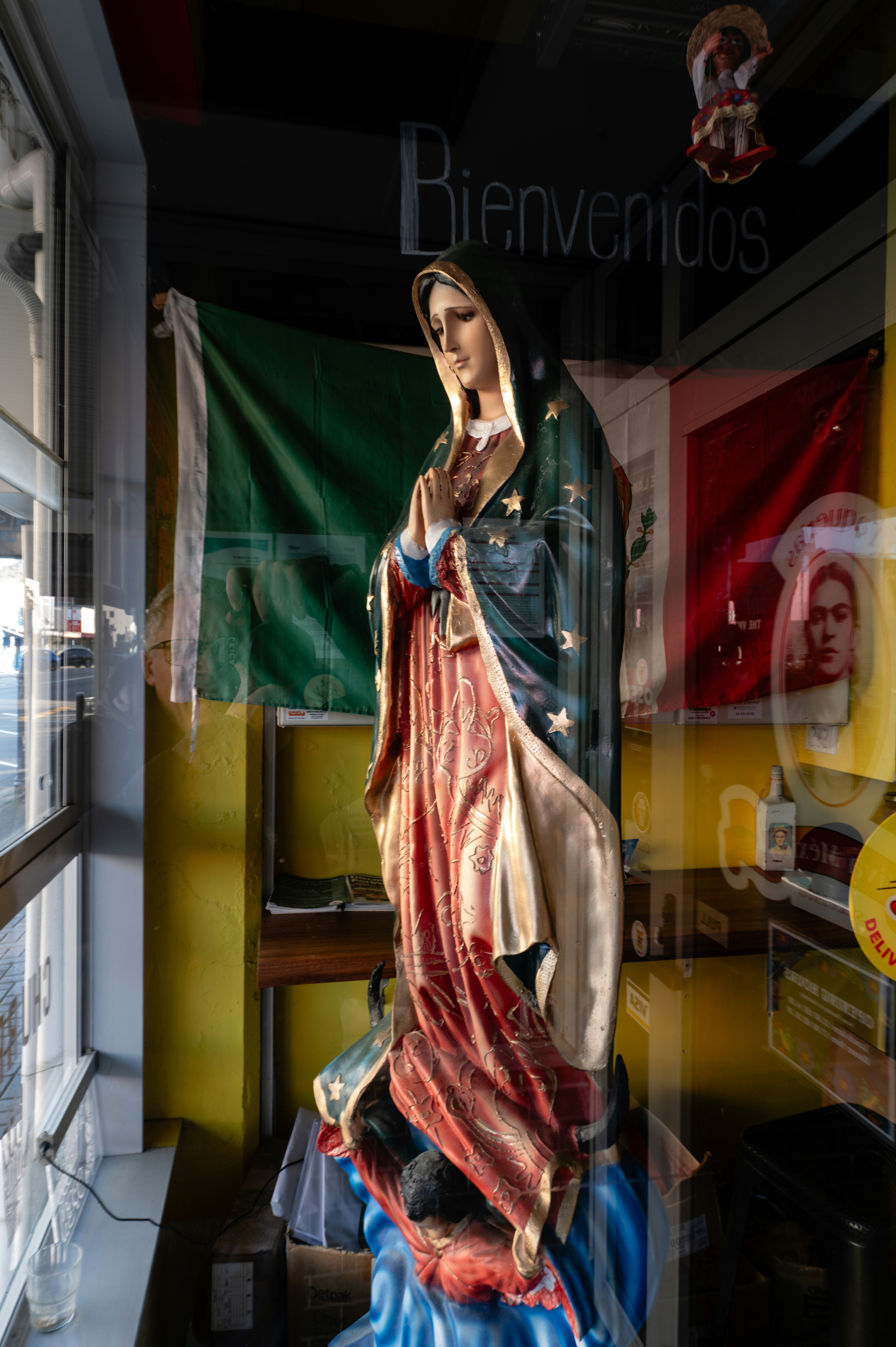 Colorful statue of a virgin Mary inside a shop window, with reflections and storefront elements creating layered depth and a busy urban backdrop.