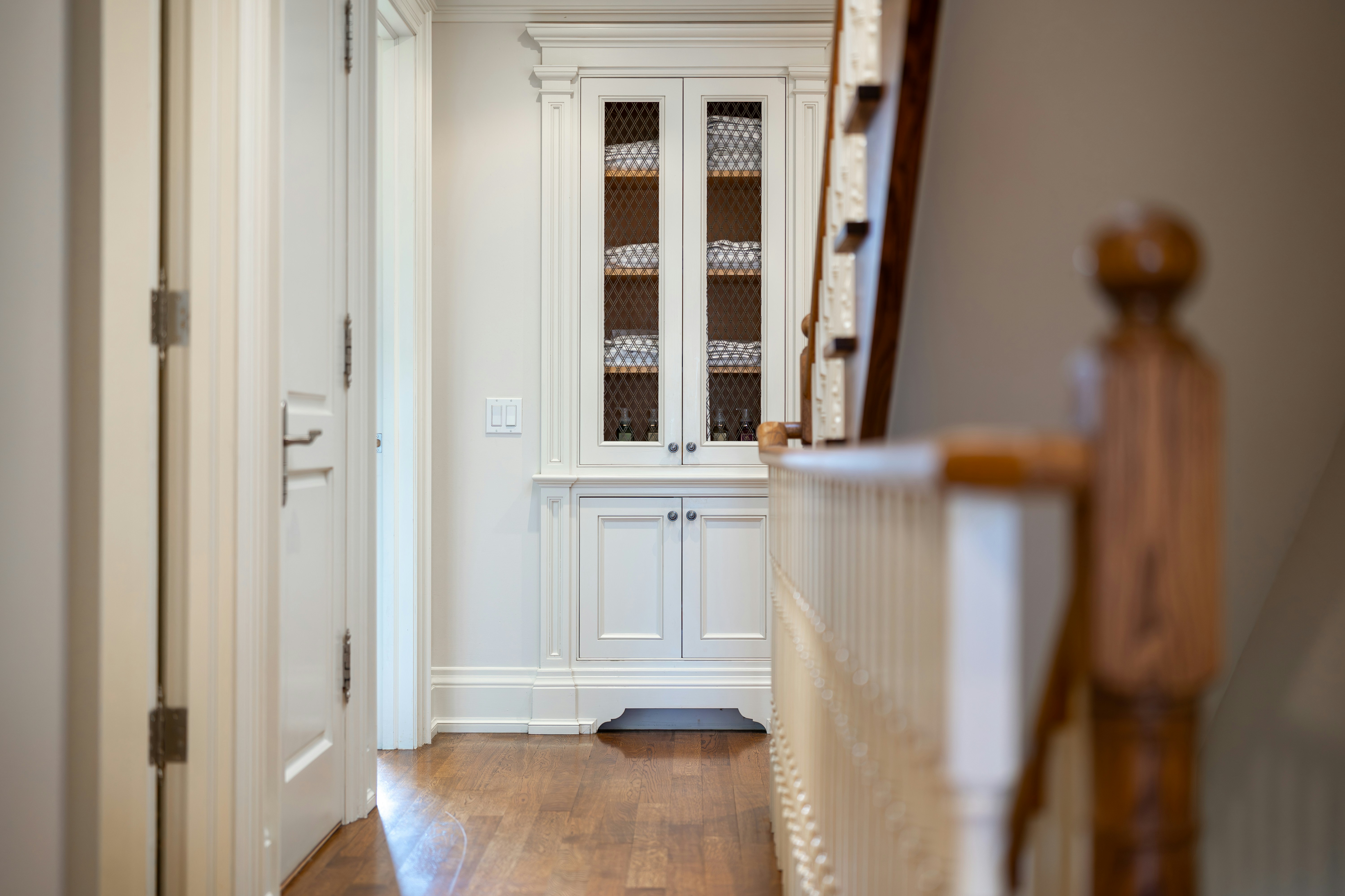 A bright, classic residential hallway featuring white built-in cabinetry, polished wood floors, white doors, and a wooden staircase railing.