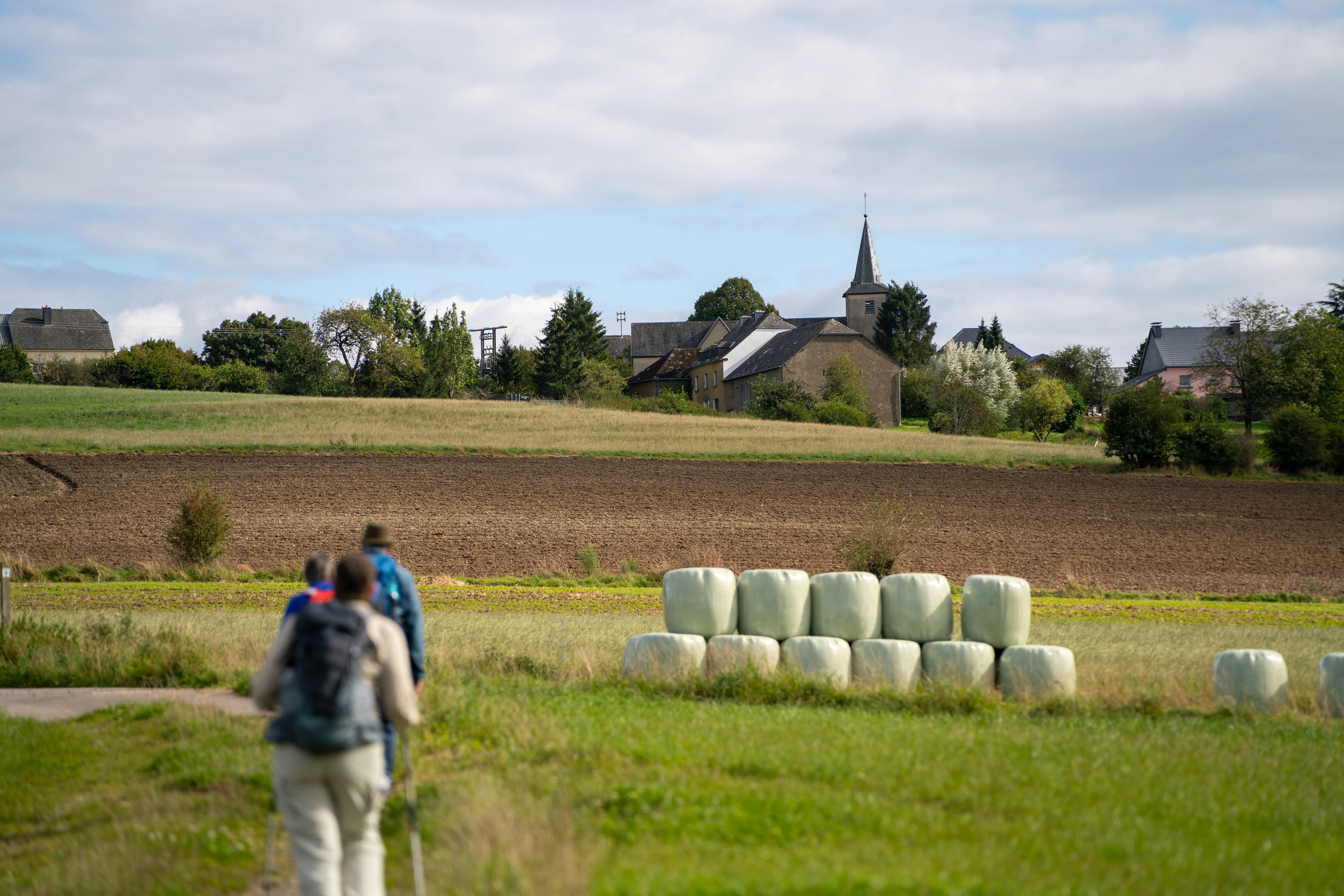 Wanderer gehen an Heuballen vorbei in Richtung eines Dorfes.