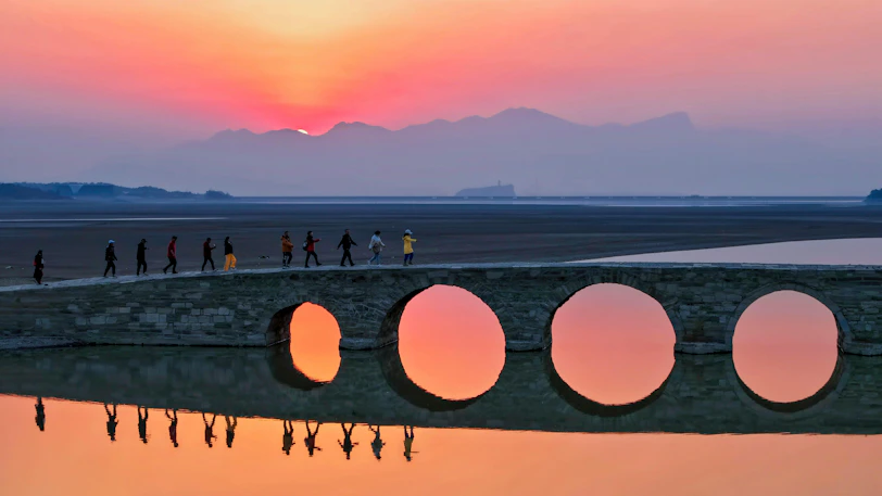 People walk across a bridge with circular openings at sunset.