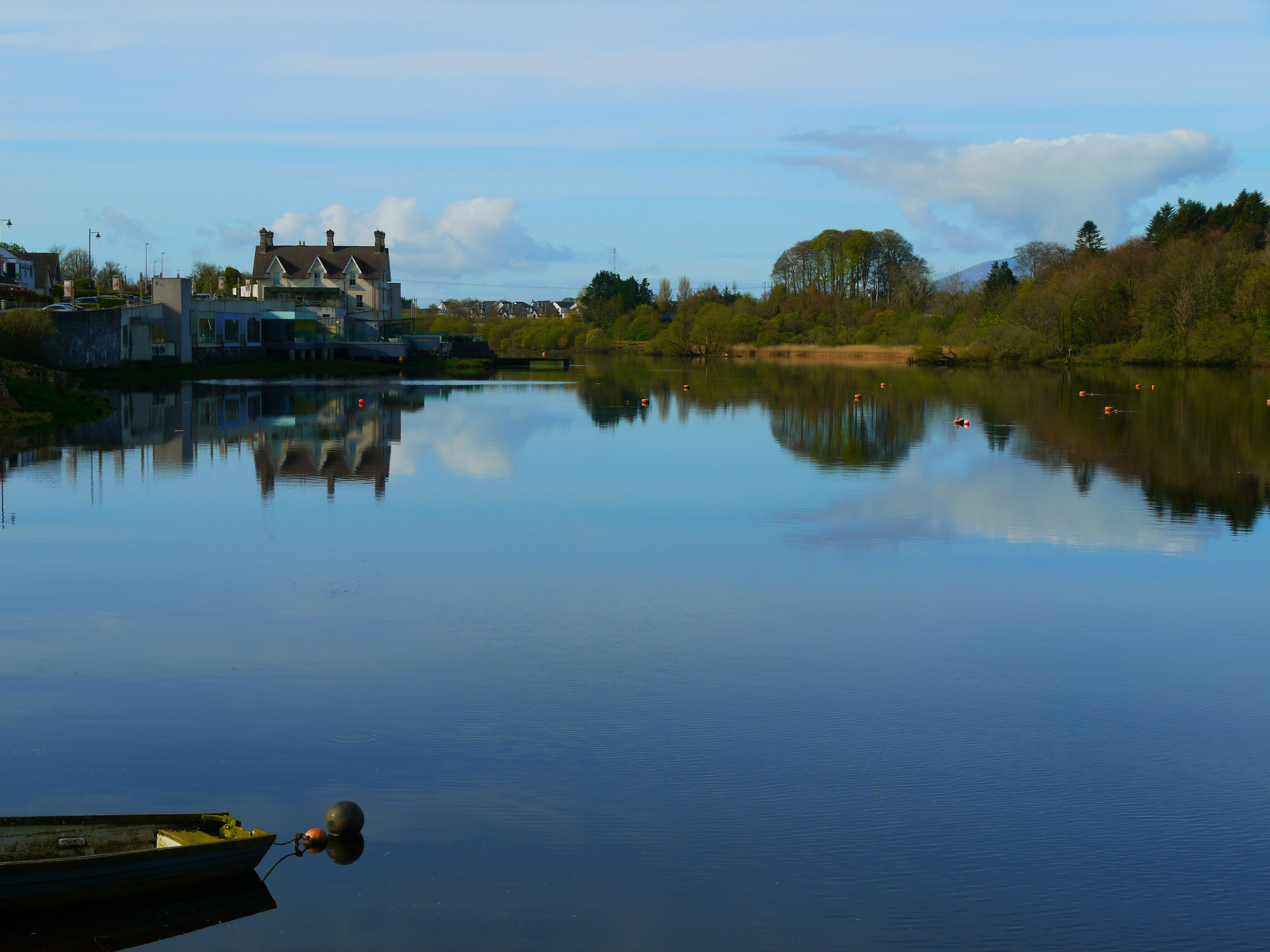 Calm lake reflects buildings and trees under blue sky.