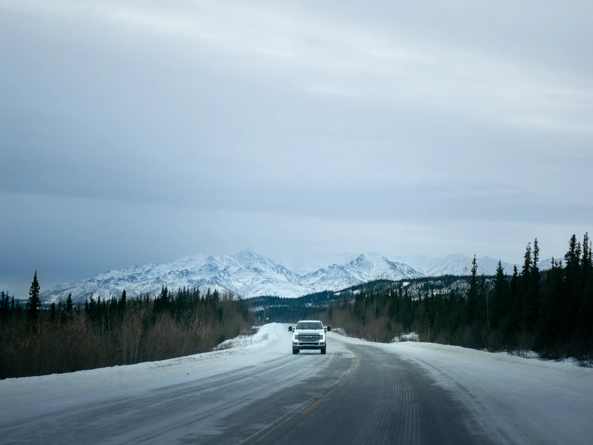A white car on a snowy road lined with spruce trees below mountains