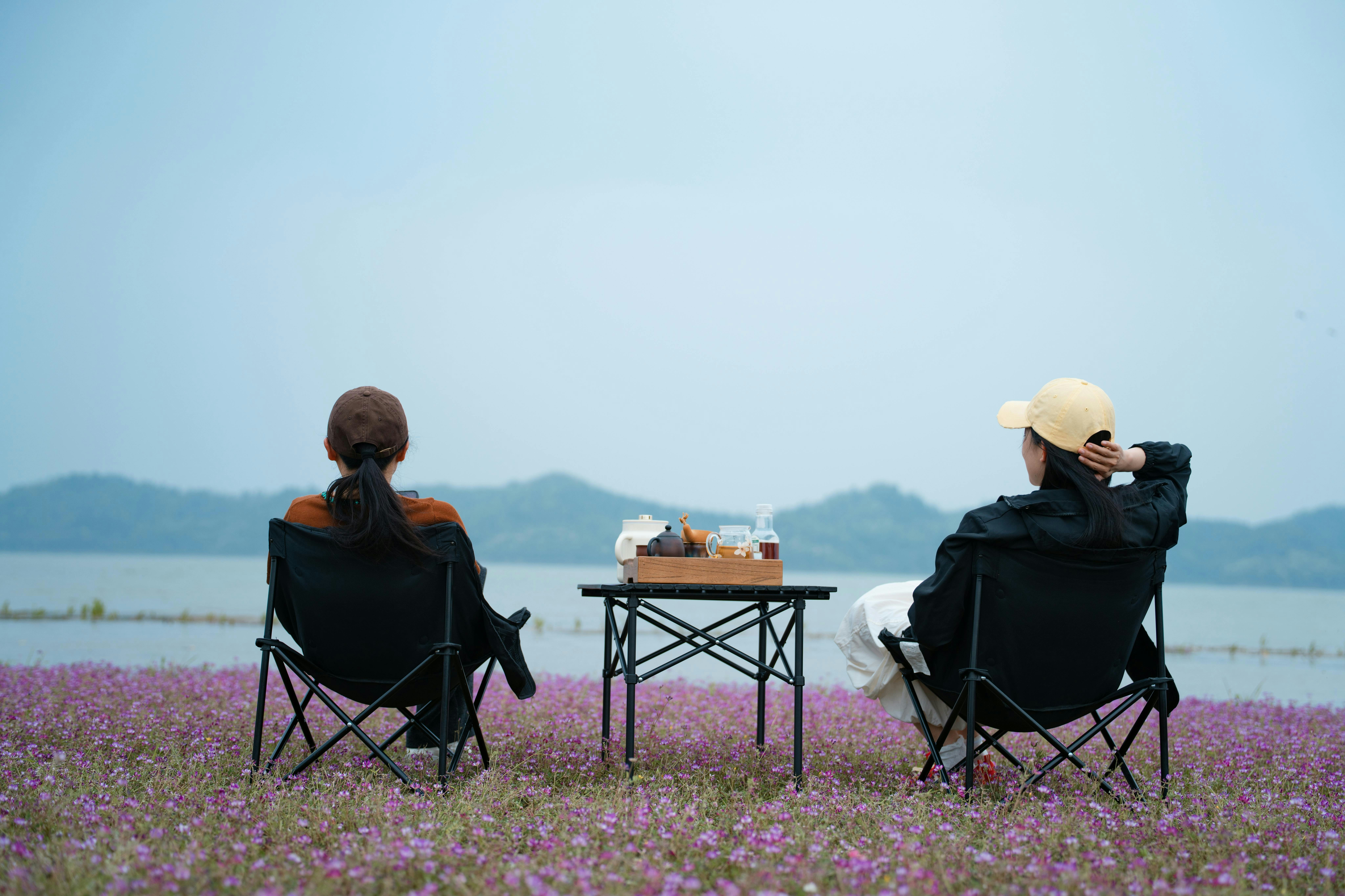 Two people in camping chairs either side of a small folding table set with a tea tray, facing a lake across a field of lilac flowers