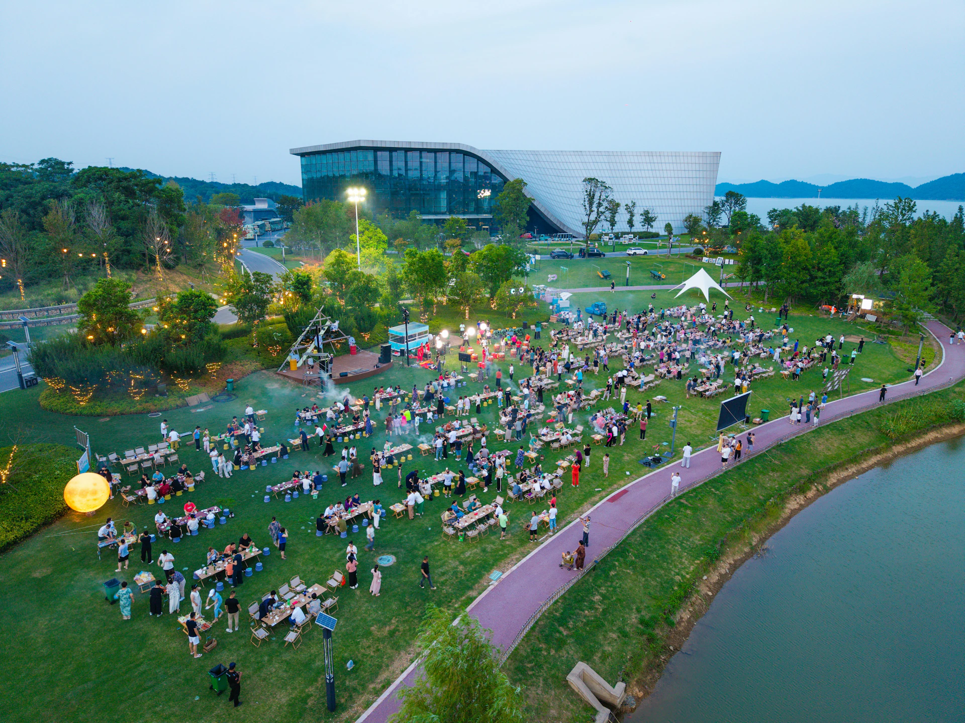 People gather on grass near a modern building at dusk.