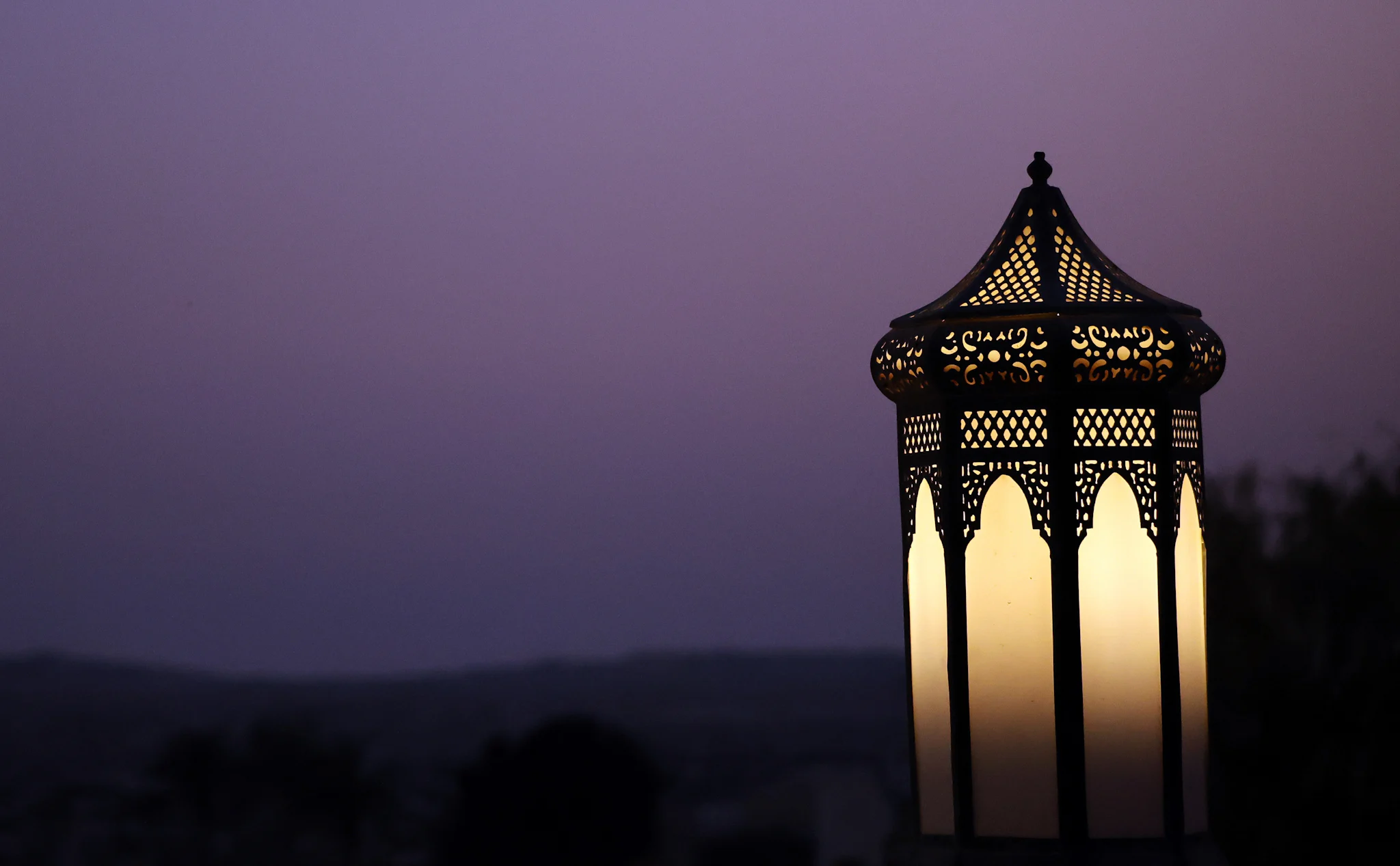 An ornate metal lantern glowing against a purple twilight sky, with dark hills on the horizon