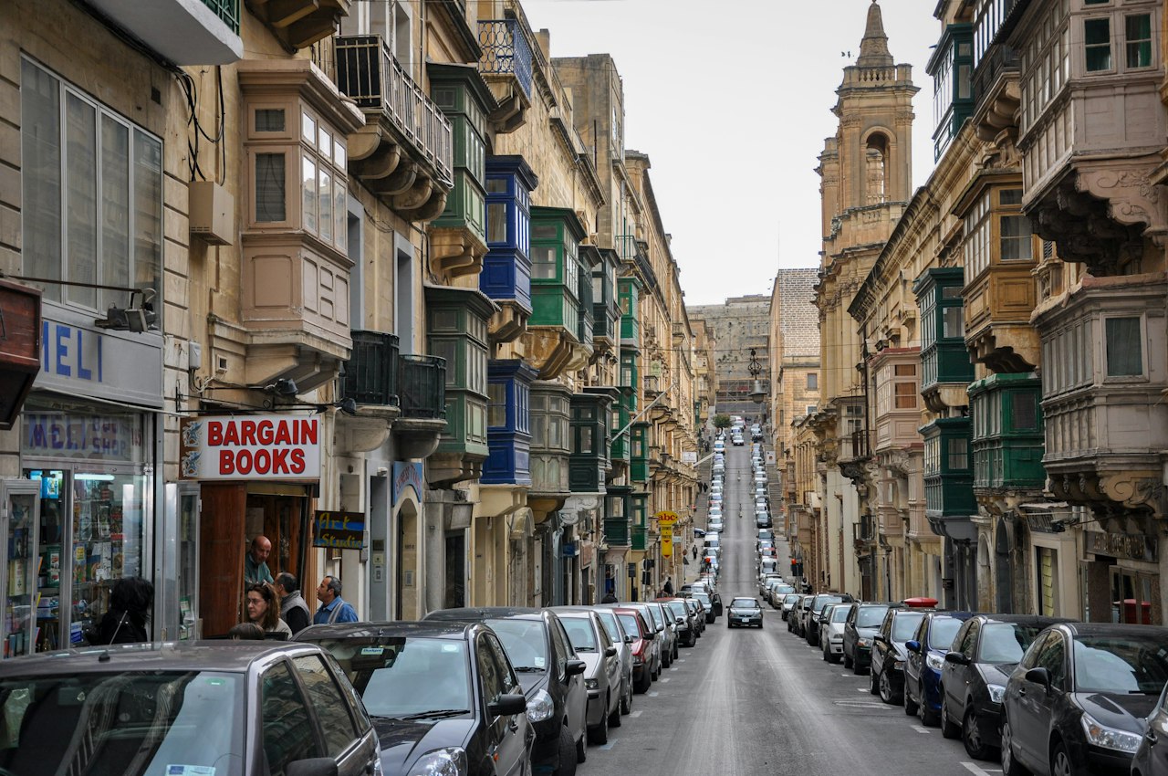 Narrow Valletta old-town street with limestone façades, Maltese balconies, parked cars, and a church tower