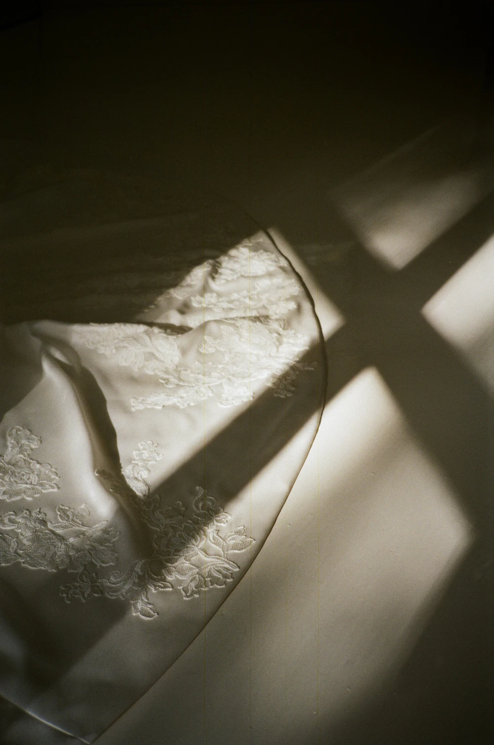 A white lace wedding dress hangs in a window, crossed by a soft shadow