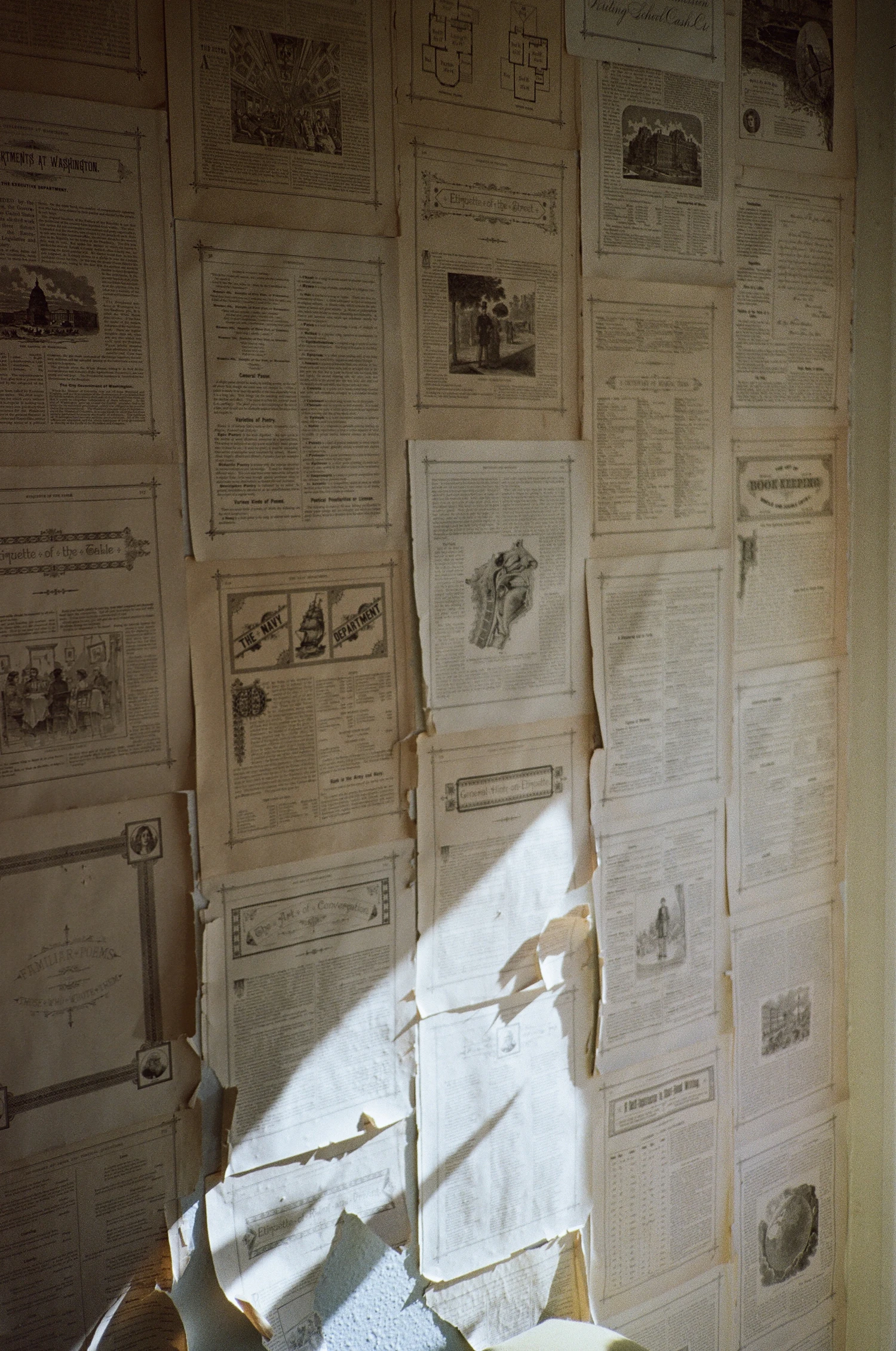A wall papered floor to ceiling with pinned printed sheets, a hard shaft of afternoon sun cutting diagonally across the lower half and tearing one page loose