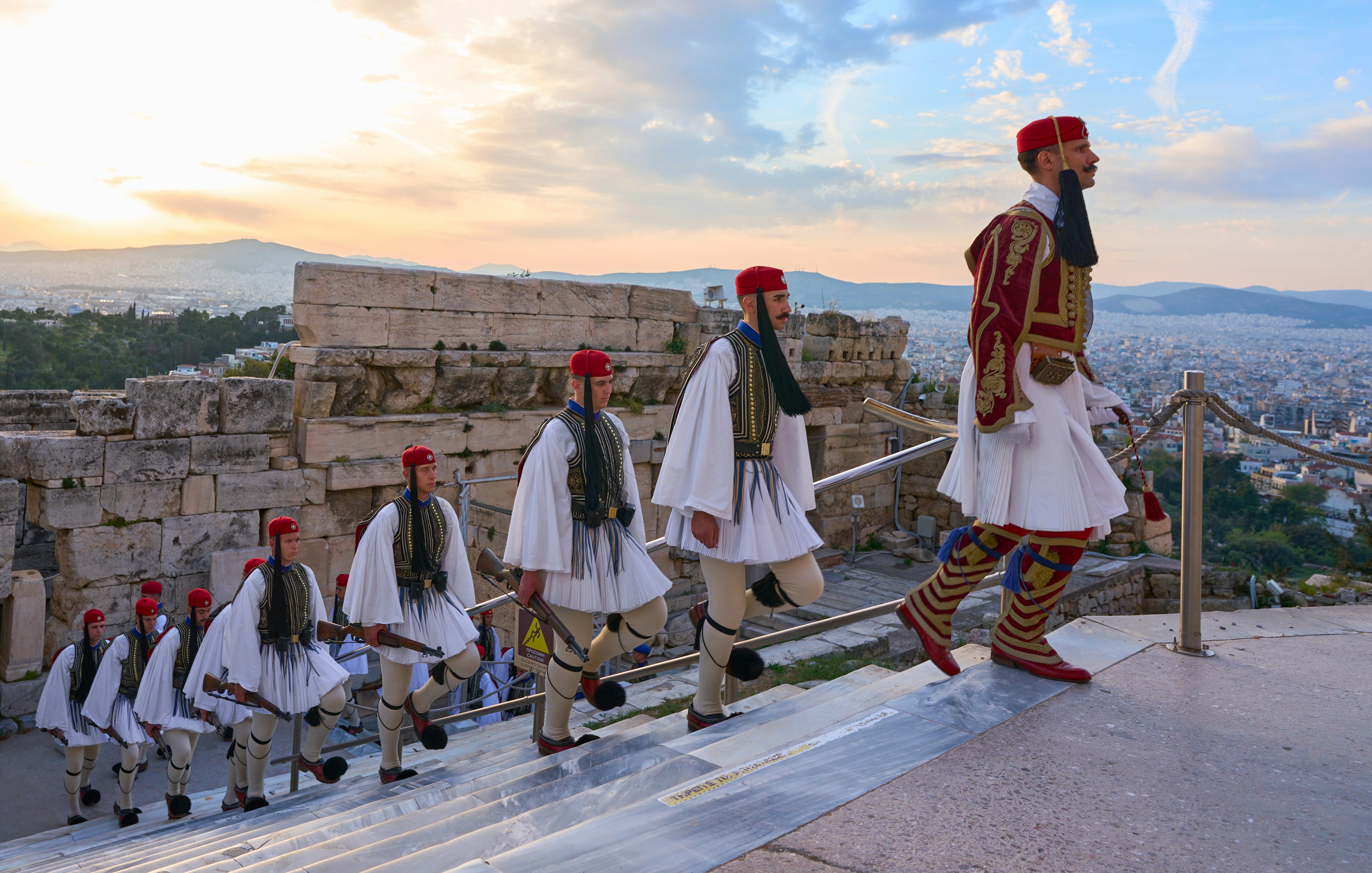 evzones in traditional uniform march up steps at acropolis